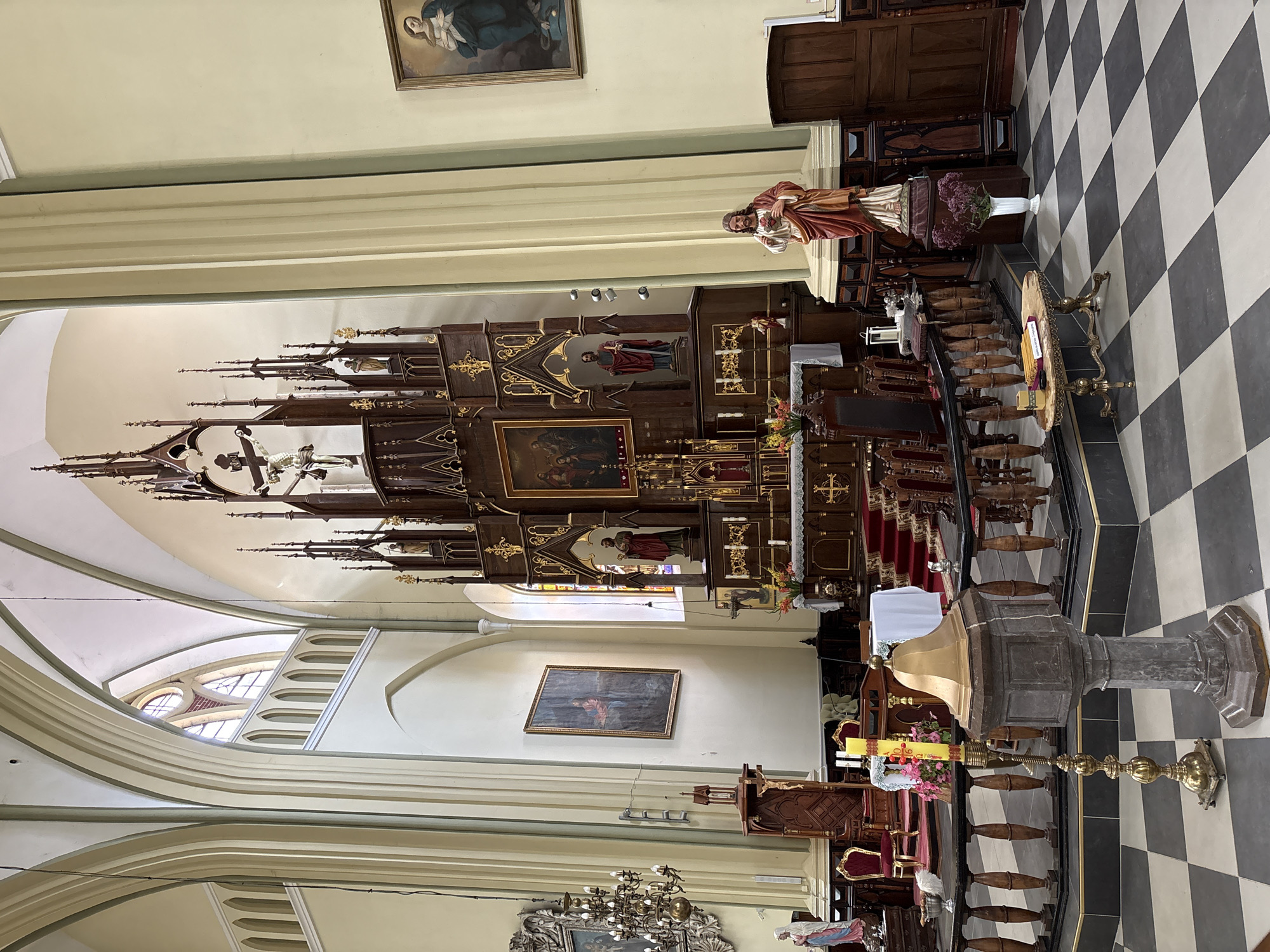 The image shows the interior of a church, likely located in Гервяцкі сельскі Савет, Belarus. The scene is dominated by a large, ornate wooden altarpiece featuring multiple levels and niches, some of which contain statues and paintings. In the foreground, a baptismal font made of stone is visible, along with a tall brass candlestick holding a yellow candle. The floor is a checkered pattern of black and white tiles. To the right of the altarpiece, a statue of Jesus with his heart exposed stands prominently. Several wooden pews are arranged on a raised platform in front of the altarpiece, some of which are covered with red carpet. The walls of the church are light-colored, with high arched windows and architectural details like ribbed vaults. Paintings hang on the walls, and decorative chandeliers are present. A statue of a kneeling woman in blue and white is visible on the left side of the image. The overall impression is one of a well-maintained, historic religious space.