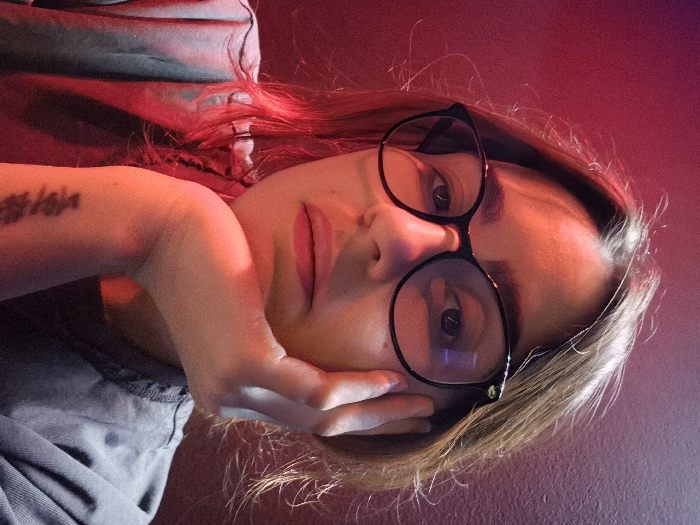 A female subject is depicted in a medium close-up photograph, indoors in Strasbourg, France. She has brown hair and wears black-framed prescription glasses. Her gaze is directed forward, towards the viewer. Her right hand supports her chin, with fingers resting along the right side of her jawline. A dark, possibly script-based, tattoo is visible on the inner aspect of her right forearm, near the wrist. She is wearing a dark, short-sleeved garment. The scene is predominantly illuminated by red light, casting a reddish hue across the subject and the plain, dark background wall.