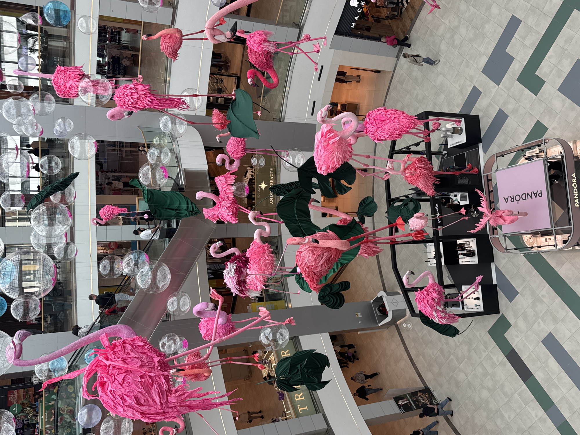 The image captures an interior view within what appears to be a shopping mall in Batumi, Georgia. The viewpoint is from above, looking down into the mall's central open area.
A prominent art installation hangs from the ceiling, featuring numerous pink flamingo sculptures interspersed with transparent globes and some large, dark green leaves. The installation is situated above various retail spaces, including a Pandora store, which is visible in the bottom right corner.
Several people are seen on different levels of the mall, walking and observing. An escalator is also visible, with people ascending and descending. The flooring design is modern with a pattern of gray, white, and green geometric shapes. The lighting is bright, suggesting a well-lit indoor environment.