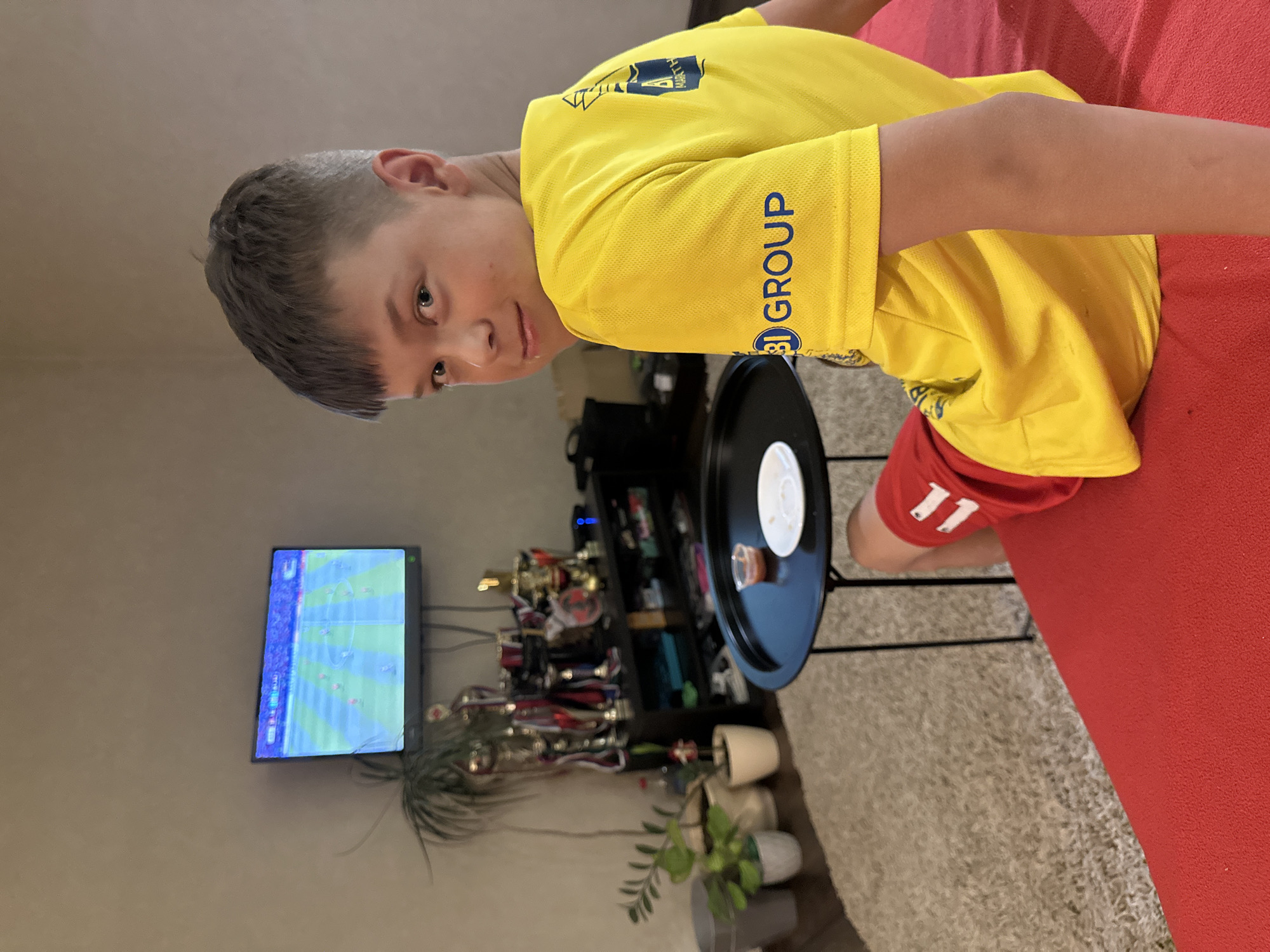 A boy wearing a yellow shirt with blue text and red shorts with the number "11" on them is lying on a red surface, possibly a couch or bed. He has short, dark hair and is looking towards the camera with a neutral expression. In the background, a television screen displays a soccer game. A shelf unit filled with trophies, plants, and other objects is visible next to the television. A black tray with a white plate and a small container is positioned in front of the boy. The setting appears to be indoors, likely a living room or playroom, with a carpeted floor. There is no indication of the time of day or weather conditions. Visible text includes "B I GROUP" on the boy's shirt sleeve and what appears to be "NORTH" above it. The number "11" is visible on his shorts.