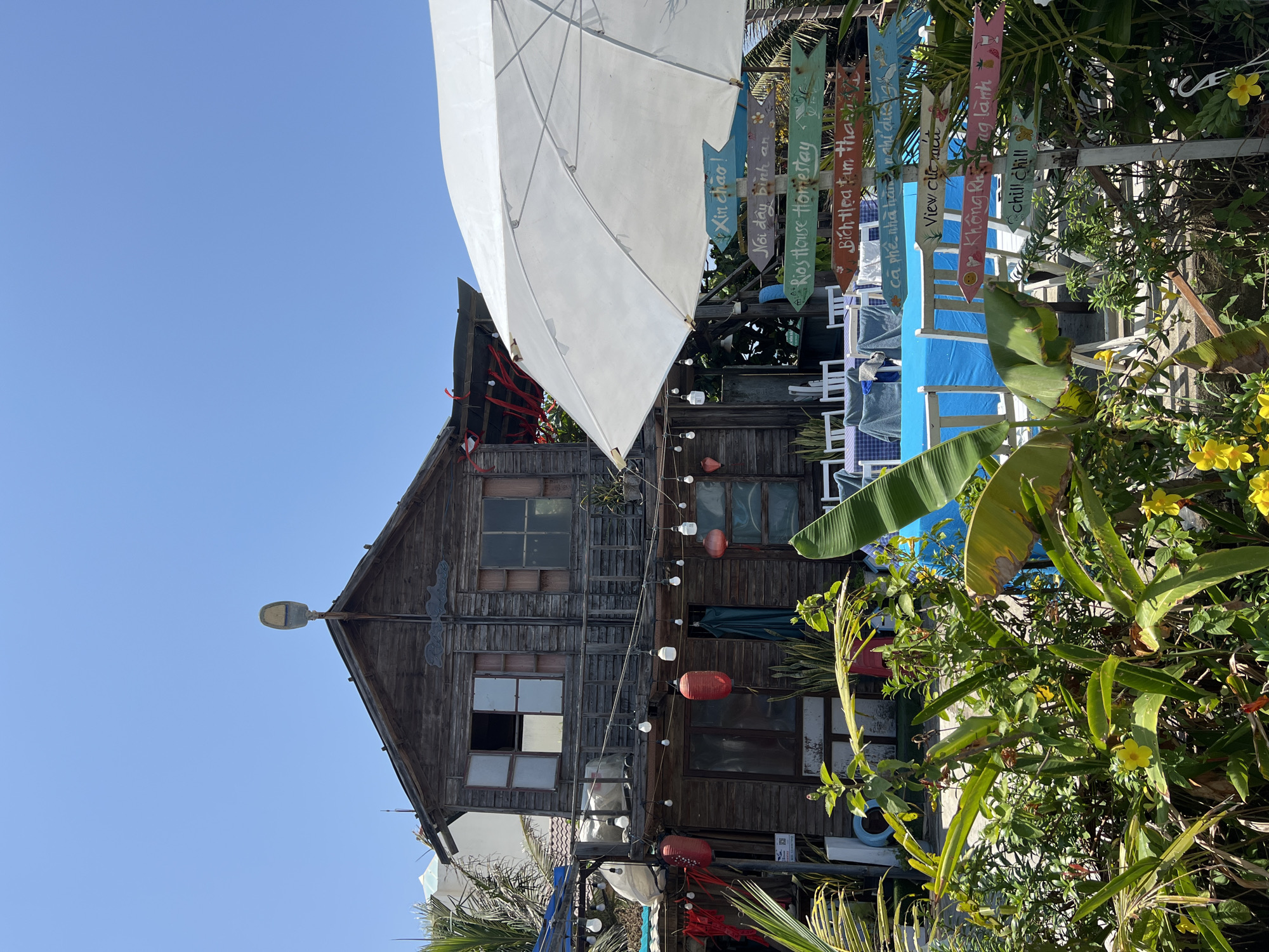 The image captures a rustic, weathered wooden building in Đà Nẵng, Vietnam, situated against a clear blue sky. The building has an A-frame roof, multiple windows, and is adorned with hanging string lights and red lanterns. A large white parasol partially obscures the building. Various plants and flowers are in the foreground, creating a dense, natural border. To the right of the building are colorful wooden signs with Vietnamese and English words. The environment has a tropical feel, suggesting a relaxed, tourist-oriented atmosphere.