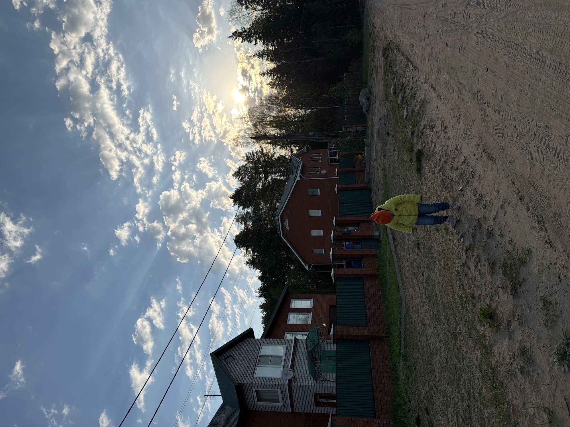 The image captures a view along a sandy path or road, adjacent to residential buildings in what is probably a suburban or rural setting. The location is stated as being in Chaikovsky, Russia. The image is oriented sideways. The sky is filled with numerous white clouds against a blue backdrop, with the sun positioned at the top of the image behind the trees. A person, wearing a yellow jacket and an orange hat, stands on the sandy ground. The residential buildings alongside the path appear to be made of brick, with green trim and fences. Trees and vegetation are visible in the background. Overhead power lines cut across the sky. The ground surface shows signs of vehicle traffic.