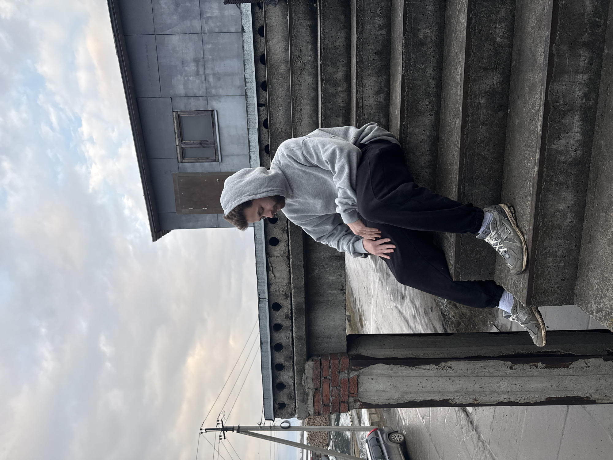 A man wearing a light gray hooded sweatshirt, dark-colored sweatpants, white socks, and gray sneakers is seen on an outdoor concrete staircase. The staircase appears to be part of a building; the man is facing down and to his left. The scene also includes part of the building's exterior, a gray sky with clouds, and a view of a nearby street including a vehicle and telephone poles. The location is the city of Chaikovsky, Russia.