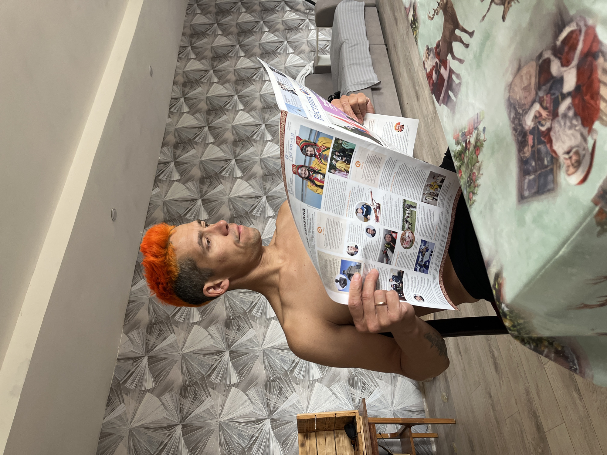 A shirtless man with orange hair is sitting at a table covered with a Christmas-themed tablecloth and reading a newspaper, "Vestnik". The room has geometric patterned wallpaper and wood flooring. The location is Chaikovsky, Russia.