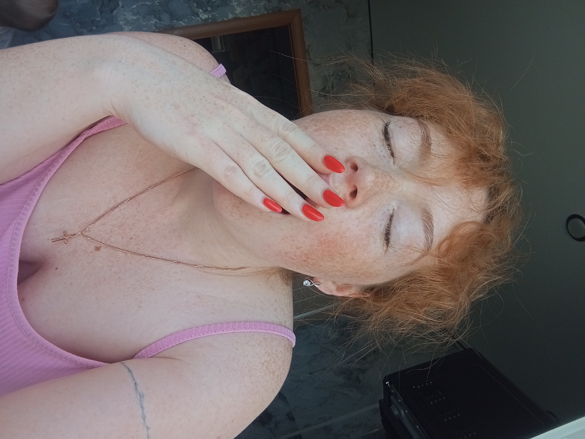 A woman with fair skin and red hair is positioned with her eyes closed, her hand covering her mouth. She wears a pink ribbed tank top, a gold chain with a cross pendant, and a silver earring in her left ear. Her fingernails are painted bright red. The background includes a dark green wall and a black object, possibly a piece of electronic equipment. There is no definitive evidence to place this scene in Yurgamysh, Russia.