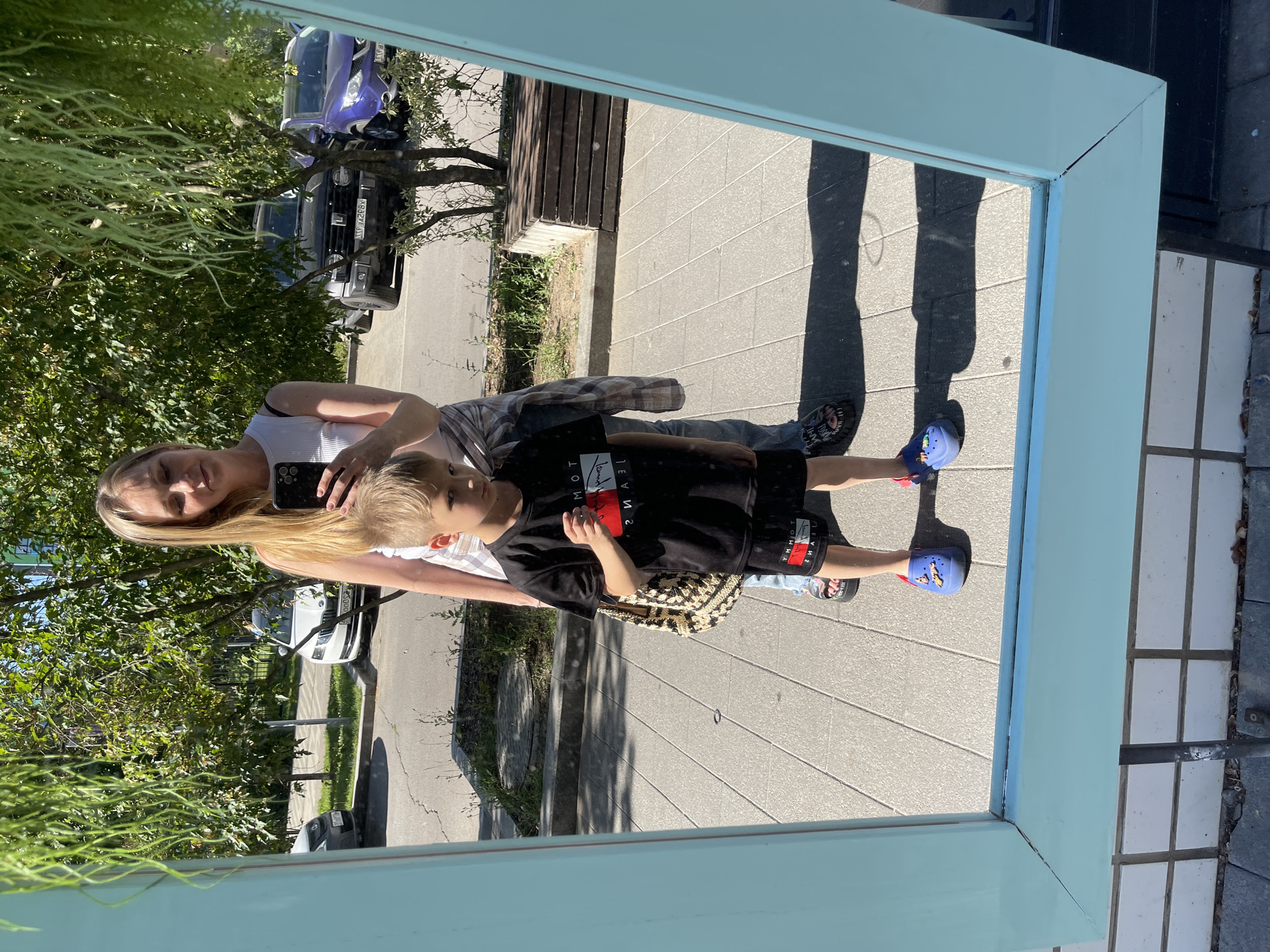 A blonde woman and a young boy are reflected in a light blue framed mirror outdoors, likely in the city of Kommunarka, Russia. The woman, wearing a white tank top and jeans, holds a black smartphone to take the photo. The boy, in a black t-shirt and shorts, stands slightly in front of her, holding a red object in his right hand. He is wearing blue Crocs, and the woman is wearing black sandals. Behind them, a street scene is visible in the mirror's reflection, including parked cars, trees, and a wooden bench. The ground is paved with grey square tiles.