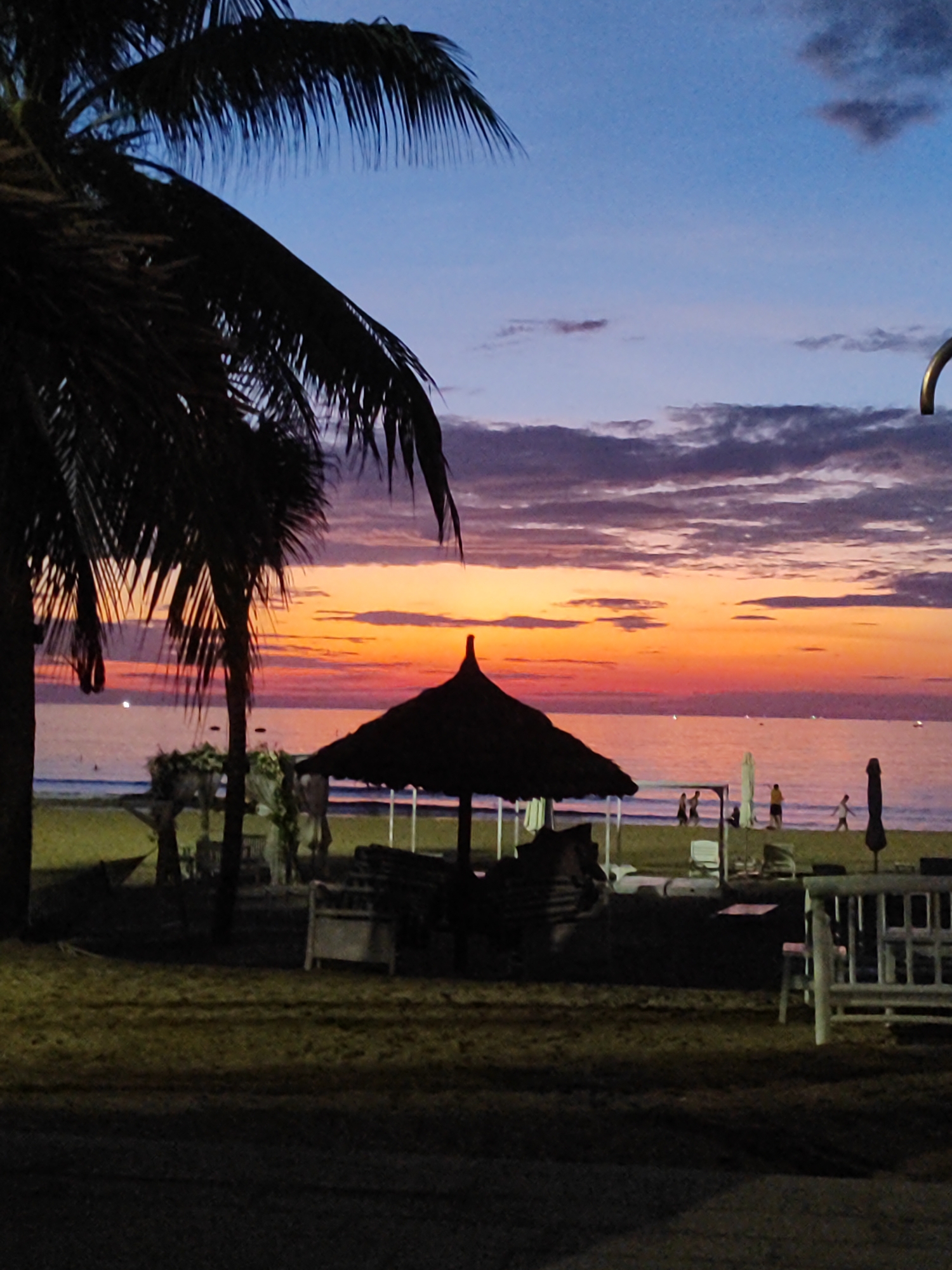 This image captures a tropical beach scene at sunset or early twilight in Đà Nẵng, Vietnam. The sky dominates the upper portion, exhibiting a gradient from deep blue at the top to vibrant orange, pink, and purple hues at the horizon. Dark, scattered clouds are present across the blue and purplish sky, silhouetted against the brighter sunset colors. The ocean surface reflects these warm tones, displaying shades of pink and purple, with several small, bright specks visible on the water, potentially distant boats or reflections.
In the foreground and midground, a dark silhouette of a large palm tree with prominent fronds occupies the left side, extending towards the center. Another, smaller palm tree is visible further back. Several dark, thatched beach umbrellas are arranged on the beach, with one being prominent in the left-center. Beneath and around these umbrellas, dark silhouettes of beach chairs or loungers are discernible. In the mid-ground, near the water's edge, several human figures are visible as dark silhouettes. A group of 2-3 individuals appears to be walking along the wet sand in the mid-right, with another person further to the right.
The immediate foreground consists of dark ground or sand, indicating low light conditions. In the lower right, white-painted wooden beach furniture, possibly a bench or chairs, is partially visible. A curved, metallic structure is partially visible in the upper right corner, cropped by the frame. The overall lighting is characterized by strong backlighting from the setting sun, creating deep silhouettes of the foreground elements against the brightly colored sky and reflective ocean.