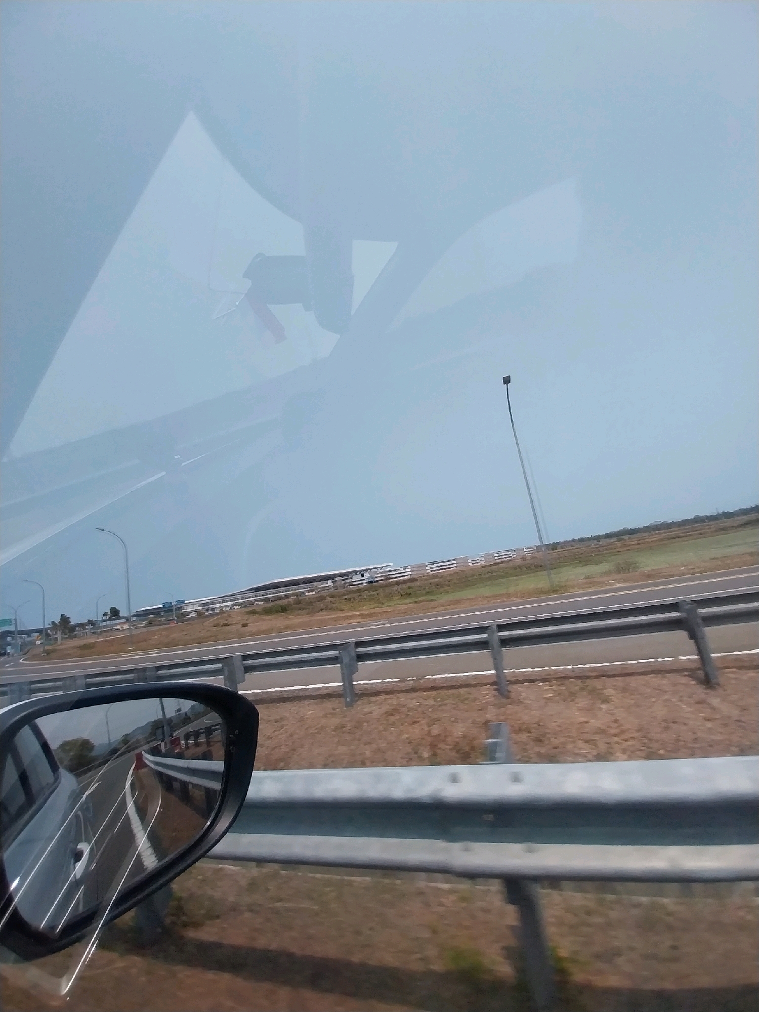 The image, taken from inside a vehicle on a highway, shows a reflection of the road and sky in the side mirror. The highway has multiple lanes, separated by a metal guardrail. In the distance, there are buildings with a flat, white roof and some vegetation. A tall, slender lamp post stands in a field beside the highway. The sky is overcast with a light blue hue. The scene is consistent with an airport perimeter in the city of Glagah, Indonesia, possibly near the Yogyakarta International Airport.