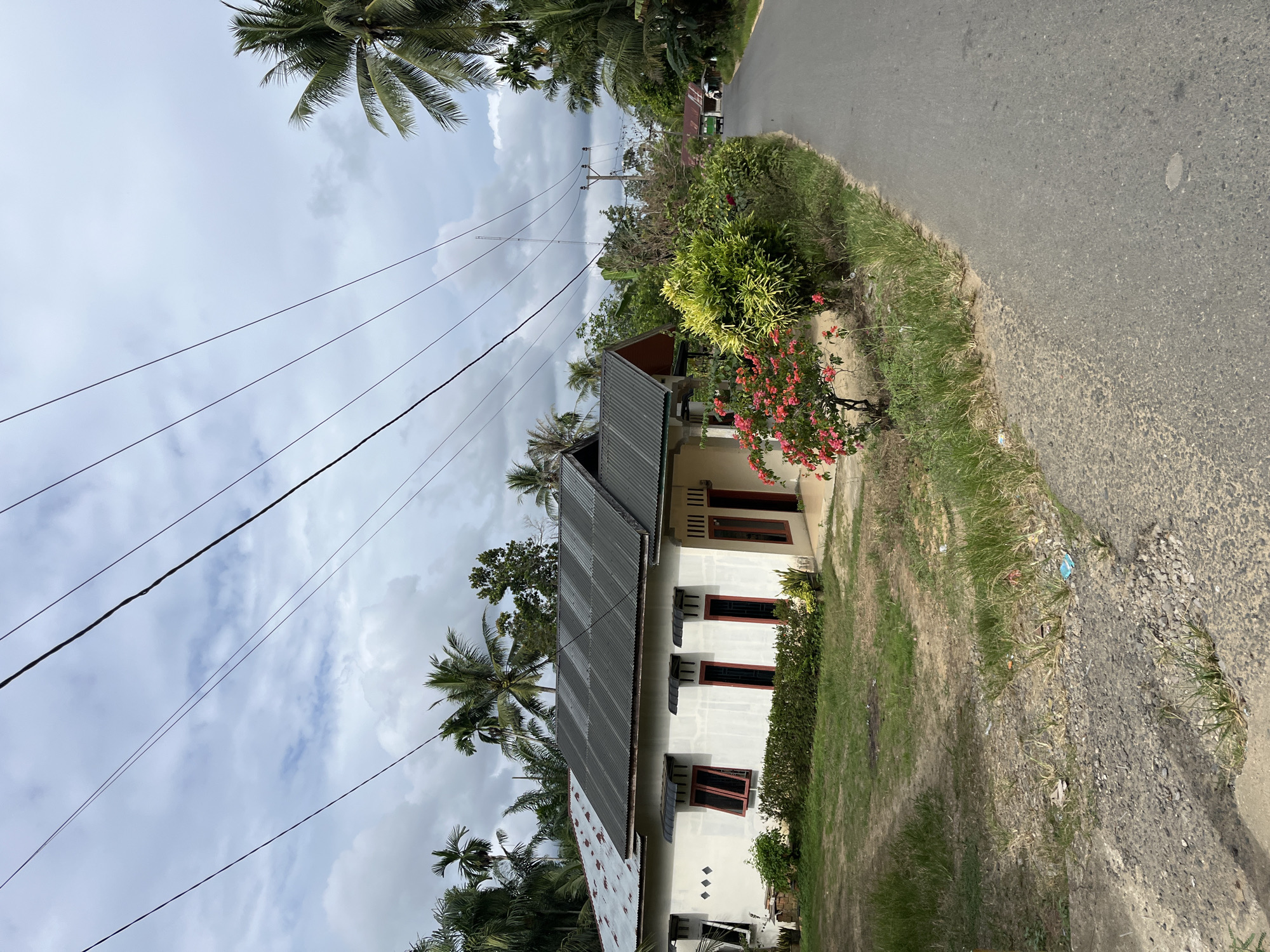 A low-angle, eye-level exterior shot captures a residential street scene under a cloudy sky in Sukajadi, Indonesia. The foreground features a paved road with sparse vegetation and gravel on its right side. To the left, a modest, single-story house with a white facade and a dark metal roof is visible. The house has several red-trimmed windows and a small porch. In front of the house, a vibrant pink flowering bush adds a splash of color. Beyond the house, palm trees and other lush green foliage dominate the background, suggesting a tropical environment. Overhead, thick black power lines crisscross the frame. The road recedes into the distance, where other houses and more vegetation can be seen. A green vehicle is partially visible on the road in the mid-ground. The overall impression is one of a rural or semi-rural residential area within a tropical climate.