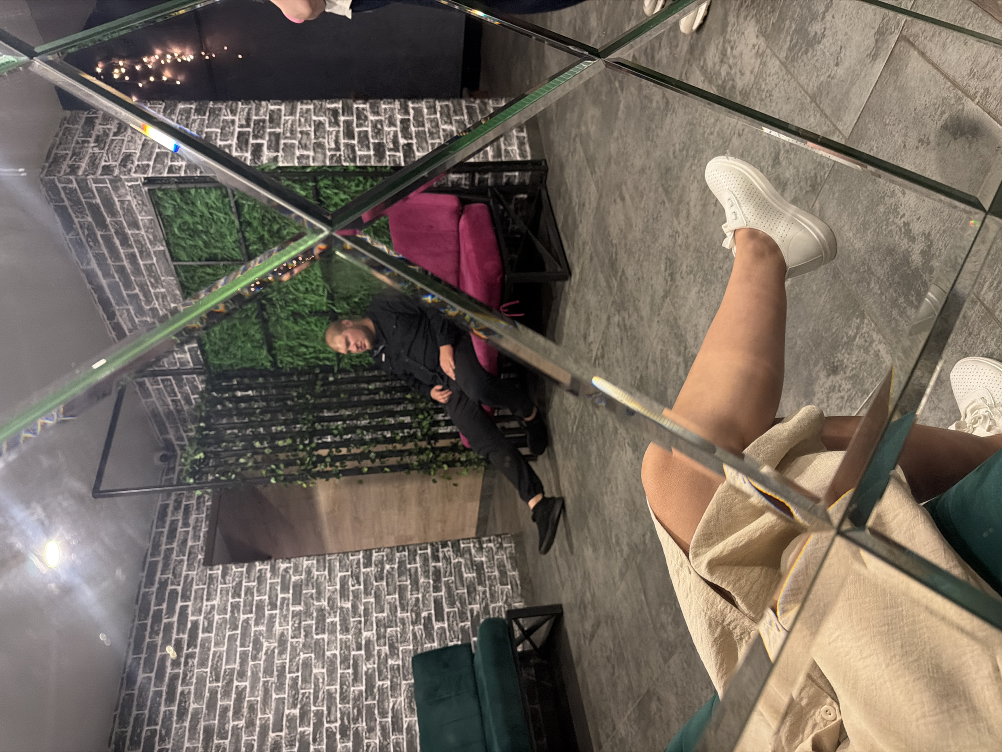 The image depicts an indoor scene with a man sitting on a magenta chair. He is wearing a black shirt and black pants. To his left, there is a dark green sofa. The walls are made of brick and there are vertical green plant decorations. The floor is tiled. A large, multi-paneled mirror is positioned in the foreground, reflecting parts of the room and two individuals, one of whom is seated and appears to be wearing a light-colored garment and white sneakers. The other reflected person is partially visible on the right side of the image, wearing white sneakers. The setting appears to be a lounge or waiting area. There is no information indicating the location within Chaikovsky, Russia.