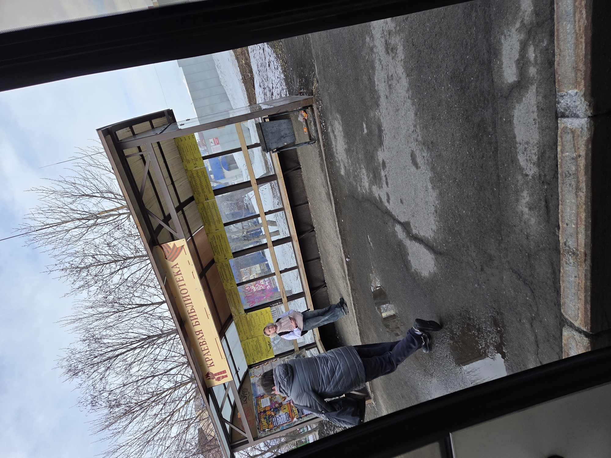 A bus stop is seen from inside a vehicle, with two people waiting. One person, wearing a grey coat, stands near the front of the bus stop. Another individual, wearing a white shirt, tan vest, and jeans, is seen seated inside the shelter. The bus stop has glass panels and displays advertisements and announcements. The area around the bus stop shows signs of melting snow, with wet pavement and puddles. Above the shelter, bare trees reach toward a partially cloudy sky. The sign above the bus shelter says "Krayeva Biblioteka" in Russian.