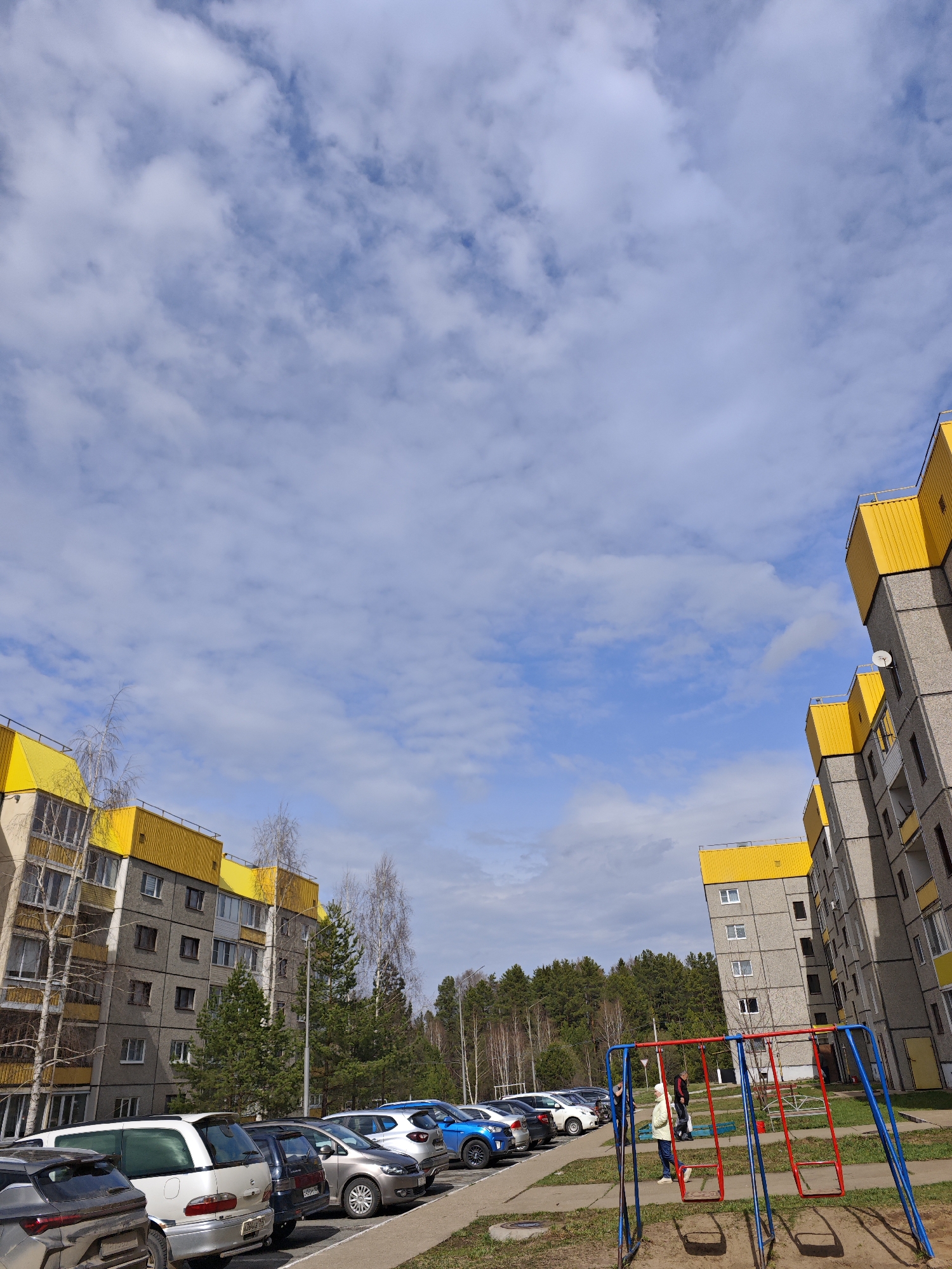 The image depicts a courtyard in Марковский, Russia, flanked by two grey apartment buildings with distinctive yellow rooftops. Parked cars line the pavement in front of the buildings. A playground with red and blue swings stands in the foreground. Two people are present at the playground. The sky is partially cloudy with patches of blue. In the distance, a line of trees is visible.