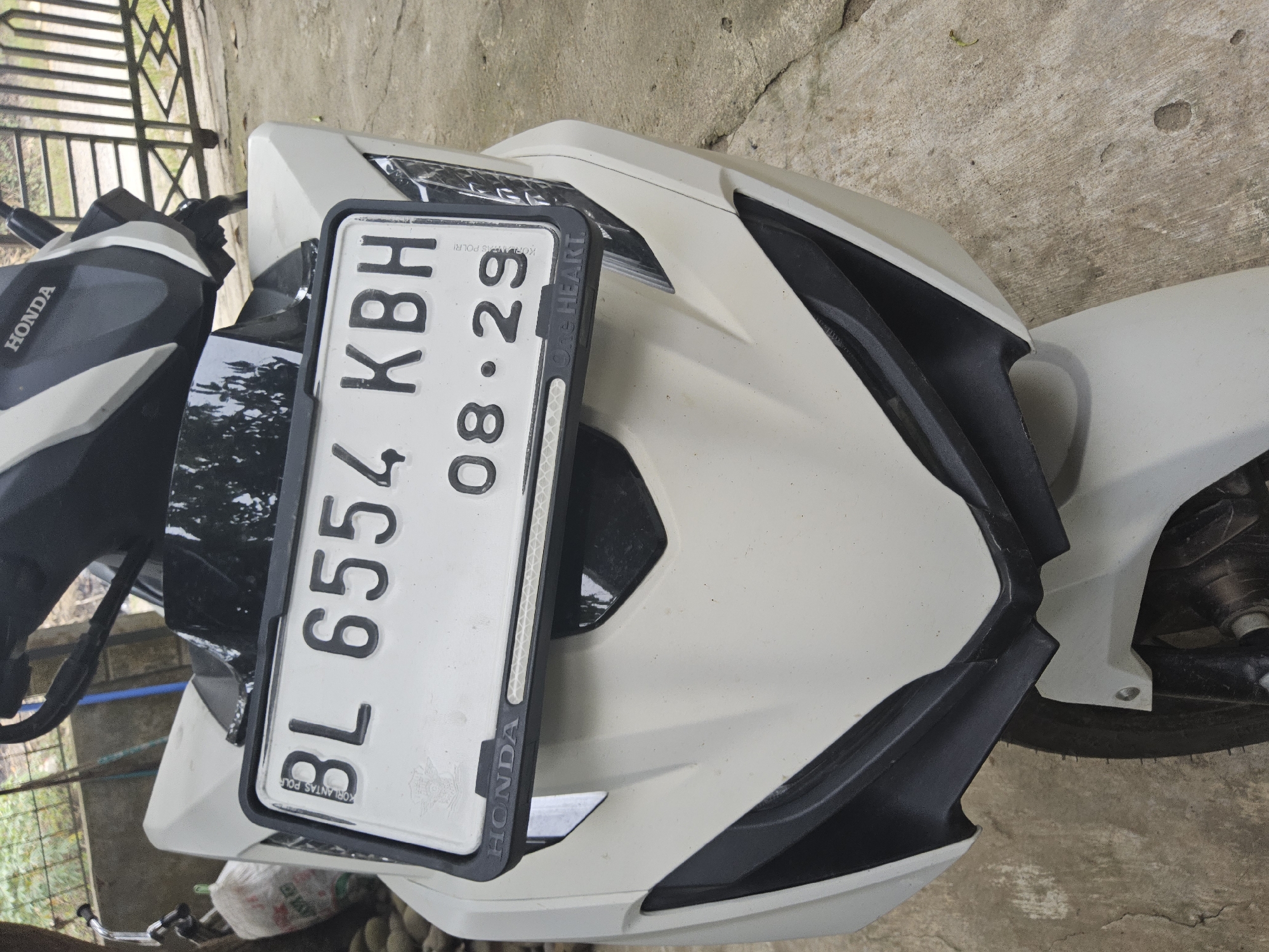 A white Honda motorcycle is parked outdoors on a concrete surface. The motorcycle has a black and white license plate with the text "BL 6554 KBH" and "08 . 29". The word "HONDA" is visible on the front of the motorcycle. In the background, a metal gate and some foliage are present. The scene appears to be located in the city of Payagaboh, Indonesia, as suggested by the license plate format. There are no people or actions depicted in the image.
