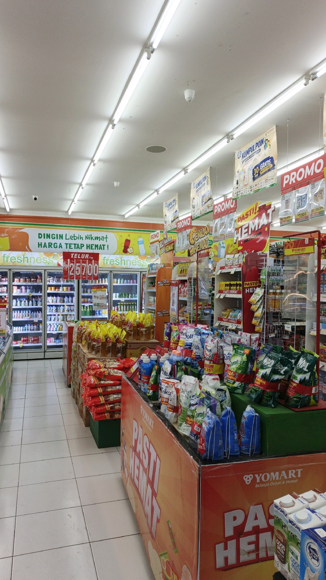 This is a photograph taken inside a Yomart convenience store in Parigi, Indonesia. The store appears to be well-stocked with various products, arranged on shelves and in refrigerated display cases. The lighting is bright, provided by fluorescent tubes on the ceiling.
In the foreground, a large promotional display stands prominently. It is red and orange, with text that reads "PASTI HEMAT" (which translates to "Definitely Save") and "YOMART Belanja Dekat & Hemat" ("YOMART Shop Near & Save"). On top of this display, several products are arranged, including bags of what appear to be snacks or laundry detergent.
To the left of the promotional display, there are refrigerators filled with beverages and other chilled items. Above these refrigerators, a banner reads "DINGIN LEBIH NIKMAT HARGA TETAP HEMAT" ("Colder is More Enjoyable, Price Remains Cheaper") and prominently displays the price "TELUR Rp 25.700" ("Eggs Rp 25.700").
Further into the store, more shelves are visible, stocked with a variety of packaged goods. Prominent red "PROMO" signs are hung from the ceiling, indicating special offers. On the right side of the image, more aisles and product displays are seen, including stacks of what look like cooking oil bottles and bags of snacks.
The floor is tiled with white square tiles. There are no people clearly visible in the photograph. The overall impression is that of a clean and organized retail environment, actively promoting sales and discounts.