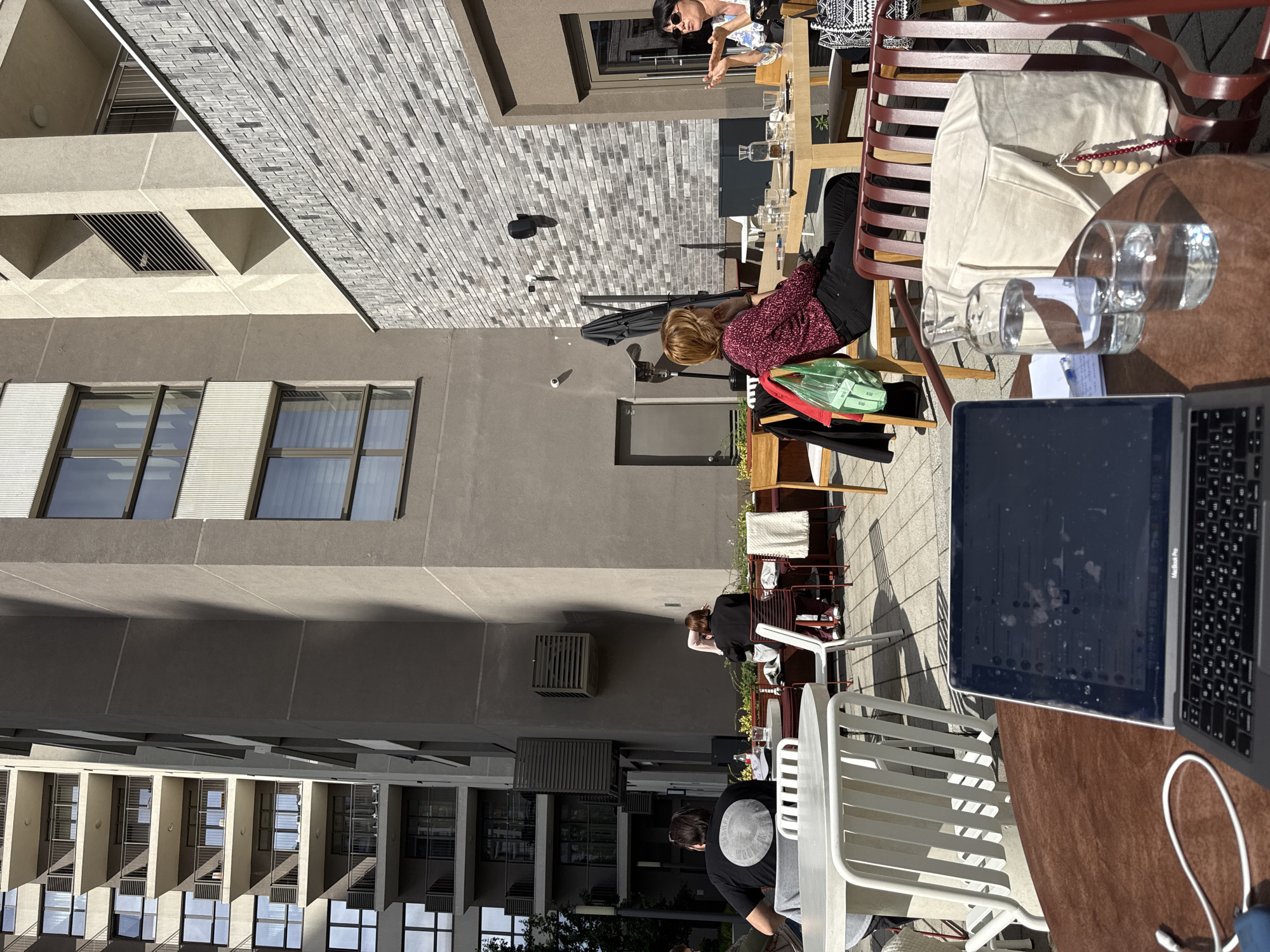 The image depicts an outdoor scene, presumably a patio or terrace, in what appears to be Belgrade, Serbia. Architectural elements of the city are visible, including buildings with multi-story window facades.
Several people are present, some interacting at tables while others are working. On the right side of the image, near the foreground, there is a laptop on a brown table next to glasses and a water bottle. A red metal chair is visible behind the table.
Further back, a woman wearing a red patterned shirt and dark pants is sitting in a wooden chair, and there's a green bag next to her. At another table, a woman wearing a white top with a black design is seated, possibly in conversation with another individual.
Further into the frame, several people are sitting in chairs, including one wearing a white hat and a black shirt that has a design on the back. The patio is furnished with a mix of wooden and metal chairs, and white plastic chairs can be seen in the mid-ground. Some plants and greenery add a touch of nature to the area. The scene is bathed in sunlight.