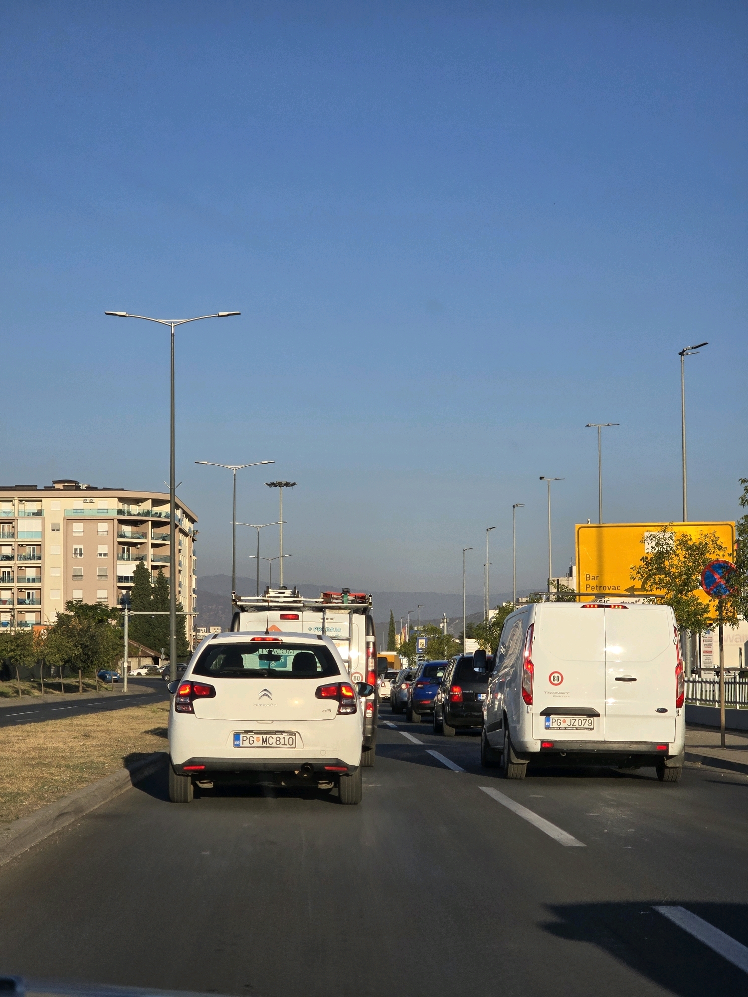 The image depicts a traffic jam on a sunny day in Podgorica, Montenegro. The perspective is from a vehicle looking forward, capturing a line of cars on a multi-lane road. In the foreground, a white Citroen C3 is visible, followed by a white Ford Transit van. Beyond these vehicles, other cars are queued up, including a blue hatchback and dark SUVs.
On the left side of the road, a modern apartment building with multiple balconies and windows stands out. Adjacent to the building are trees and a grassy area. The road is lined with tall streetlights on both sides, and a large yellow directional sign indicating "Bar" and "Petrovac" is visible on the right. In the distance, mountains are faintly discernible against a clear blue sky. The overall scene is indicative of typical urban traffic flow during daylight hours.