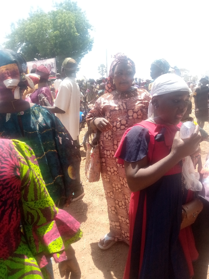 The scene depicts an outdoor public gathering under bright daylight in Kagoma, Nigeria. The ground is dry, reddish-brown earth. In the foreground, multiple individuals are present. On the far left, a person is partially visible wearing a green and pink patterned garment. To their right, an individual with their back to the camera wears a blue patterned robe and a light-colored head covering that obscures their face. Adjacent, a person in a loose white tunic and a dark head covering also faces away from the camera. In the center, a woman with a neutral expression wears a patterned brown and white long dress and matching headwrap, holding a light-colored patterned bag in her left hand. She wears white flip-flops. To her right and slightly in front, a younger individual, wearing a red and dark blue dress and a white head covering, holds a crumpled white plastic object in their right hand and has a small brown purse on their left shoulder. In the midground, a large white banner featuring images of three men and partial text, indicative of a political or community event, is visible, flanked by two microphone stands. A dense crowd of people in varied attire fills the background. A large green leafy tree is visible in the upper left background against an overexposed white sky.