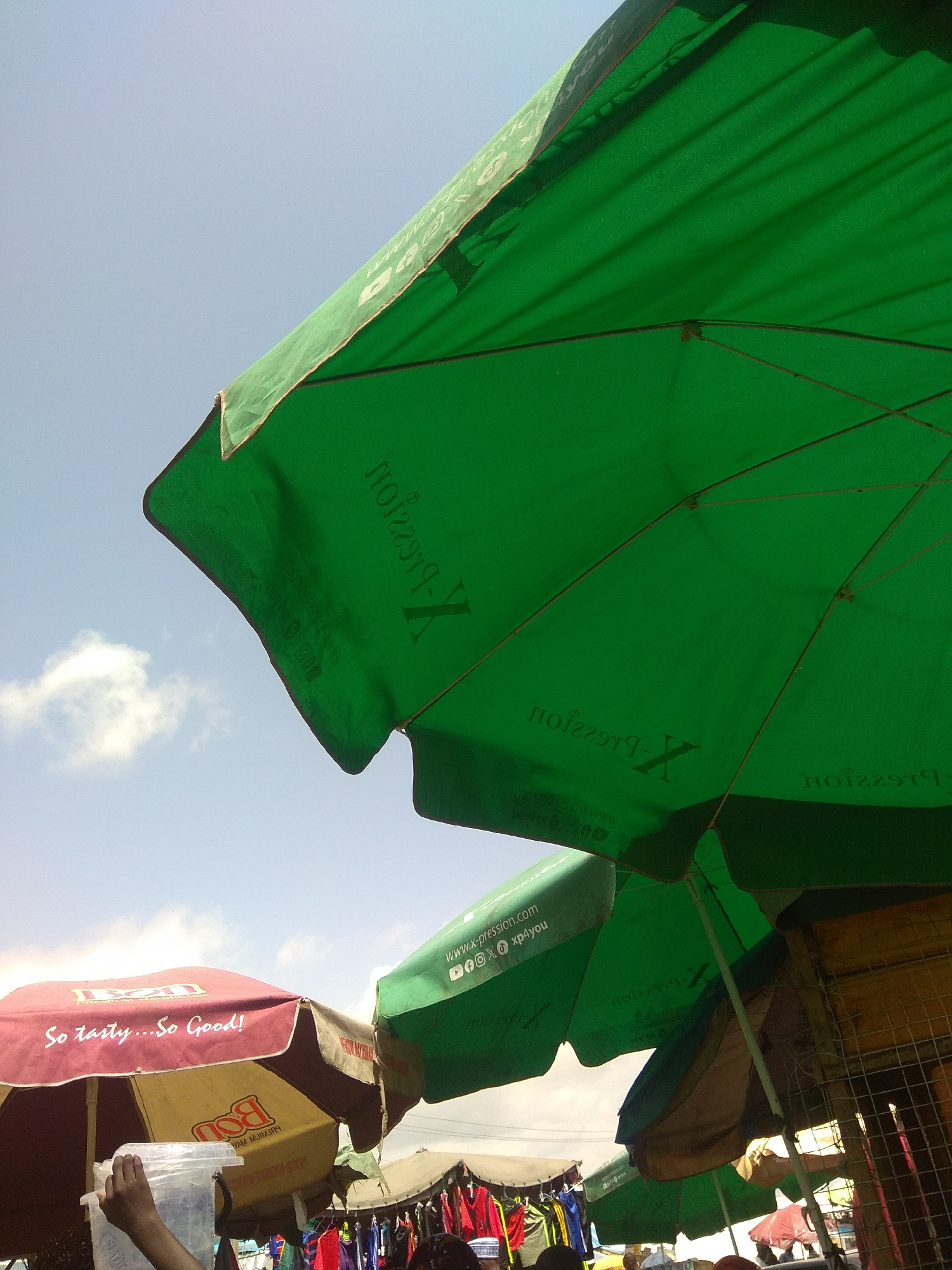 This image depicts an outdoor market scene in Jalingo, Nigeria, characterized by stalls and vendors operating under various umbrellas. The primary focus is on several large, green umbrellas, with one prominently angled towards the sky, revealing its underside and support structure. These umbrellas appear to be branded, with visible text like "X-pression" and "www.x-pression.com" on their surface.
In the background, a red and yellow umbrella is visible, advertising a product with the text "So tasty...So Good!" and "BON PREMIUM MAYONNAISE". Another umbrella, partially obscured, displays the brand "BON". The sky above is a pale blue, with a few scattered white clouds.
To the left, a person's hand is seen holding a clear plastic container. Further back, the market extends with more stalls, including racks of clothing, suggesting a vibrant commercial activity. The overall impression is a busy marketplace under the open sky, with a reliance on umbrellas for shade.