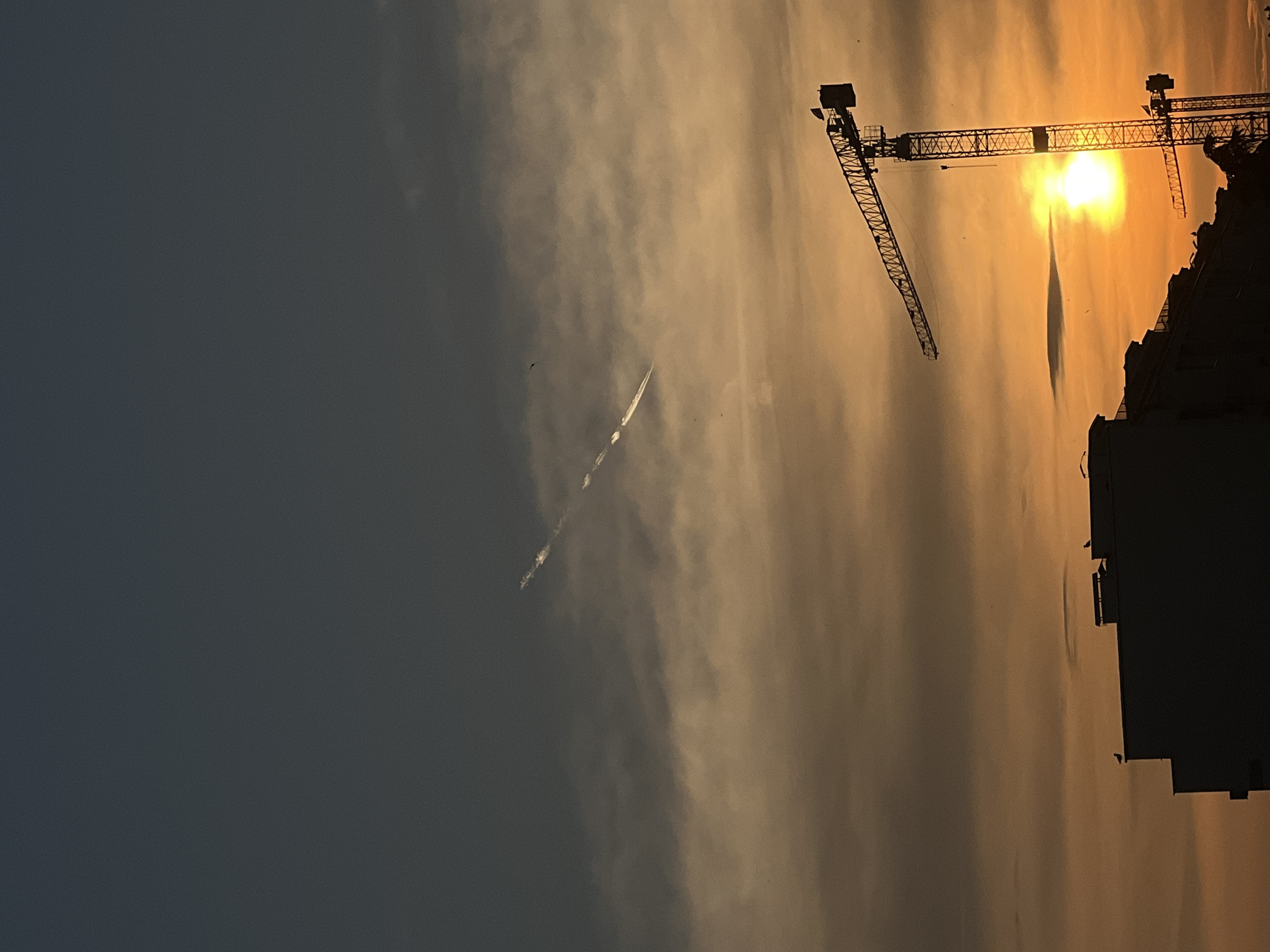 This image captures an urban sunset scene in the city of Burgas, Bulgaria. The sky is divided into distinct tonal bands: the upper portion is a deep, muted blue-grey, transitioning to warm orange and yellow hues at the horizon, illuminated by the setting sun. Faint, dissipating contrails from an aircraft are visible in the upper-middle sky, appearing as two parallel, short white streaks against the darker blue and lighter orange clouds. Scattered, indistinct dark specks, likely birds, are present in the sky.
In the foreground, the silhouettes of buildings and construction cranes dominate the lower half of the frame. On the left, a multi-story building, likely an apartment or office complex, presents a dark, featureless outline against the illuminated sky. Several small, dark shapes, identified as birds, are perched on the rooftops of these buildings. To the right, two prominent tower cranes stand silhouetted. The larger, more central crane is a lattice structure, with its horizontal jib extending towards the left and its mast rising vertically. The intense, circular disc of the setting sun, a brilliant yellow-orange, is positioned behind and slightly to the right of this crane's mast, casting a strong glare. A second, smaller tower crane is partially visible behind and to the right of the larger one. The overall scene depicts an active construction or industrial area against a dramatic twilight sky.