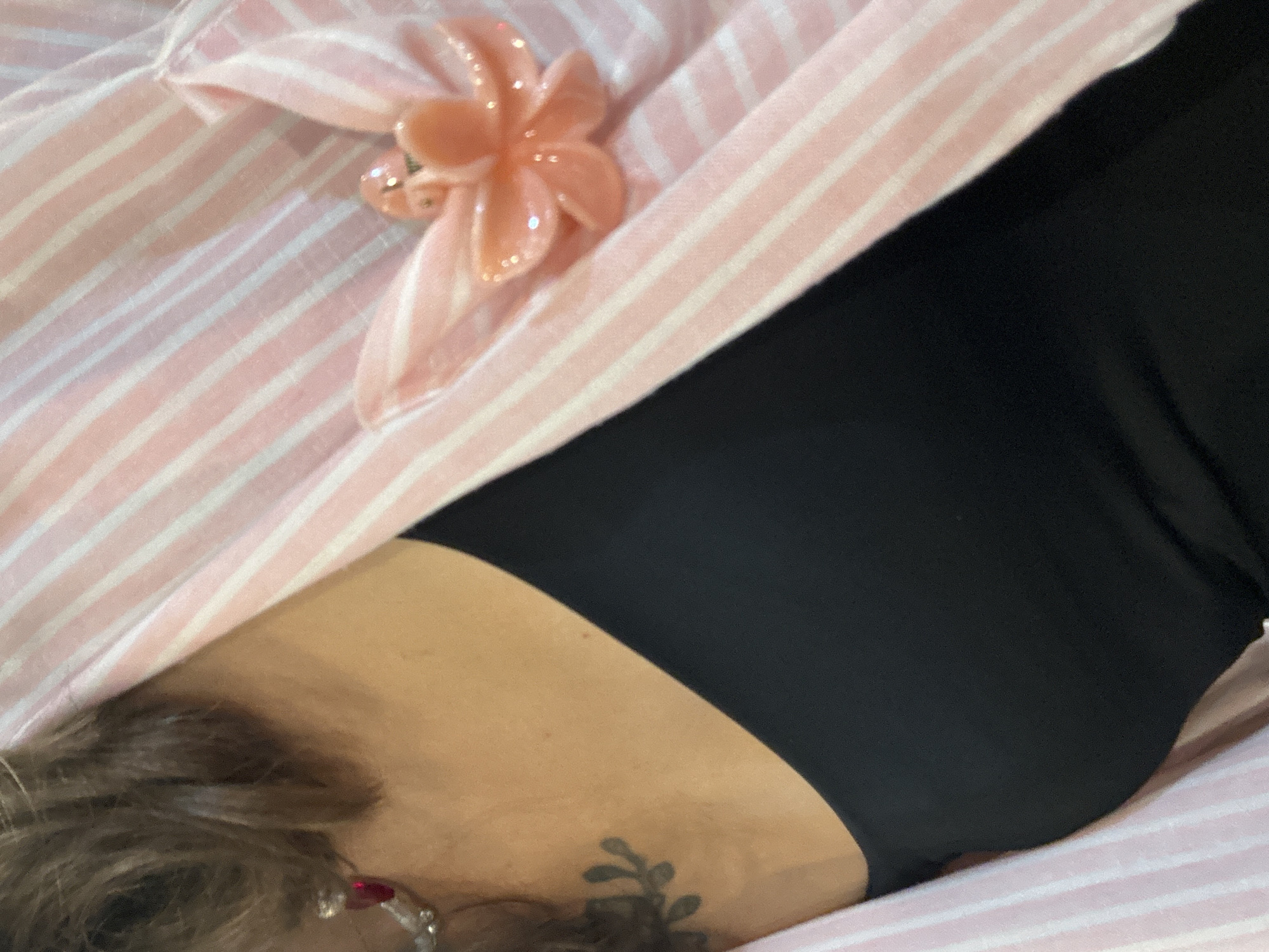 A close-up shot features a person's upper chest and neck area, adorned with jewelry and clothing. On the left side of the frame, a part of the person's shoulder and upper arm is visible, with a tattoo of a leafy branch on the skin. A silver earring with a red gem hangs from the ear. The person is wearing a black top, and over it, a pink and white striped blazer or shirt. A decorative brooch shaped like a peach-colored flower with glitter accents is pinned to the lapel of the striped garment. The lighting is soft, highlighting the textures of the fabric and the details of the jewelry. The background is blurred, suggesting a shallow depth of field. No specific location in Ratsada, Thailand, can be determined from the image.