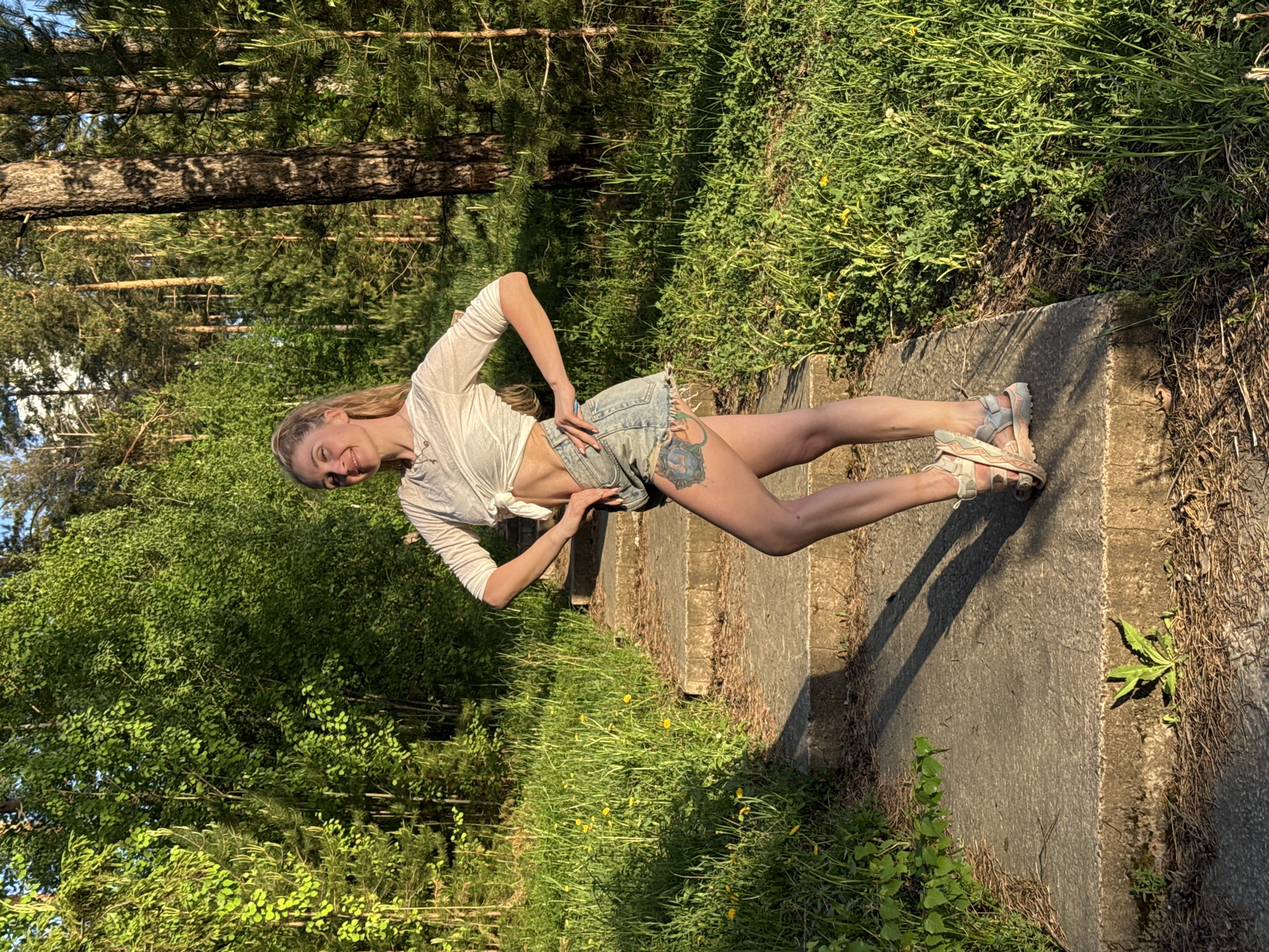 A woman with long blonde hair, wearing a white long-sleeved shirt tied at the waist, denim shorts, and sneakers, is standing on a set of concrete steps. She has a tattoo on her right thigh. The background consists of a forest setting with trees and green foliage. The location is identified as Noviy, Russia.