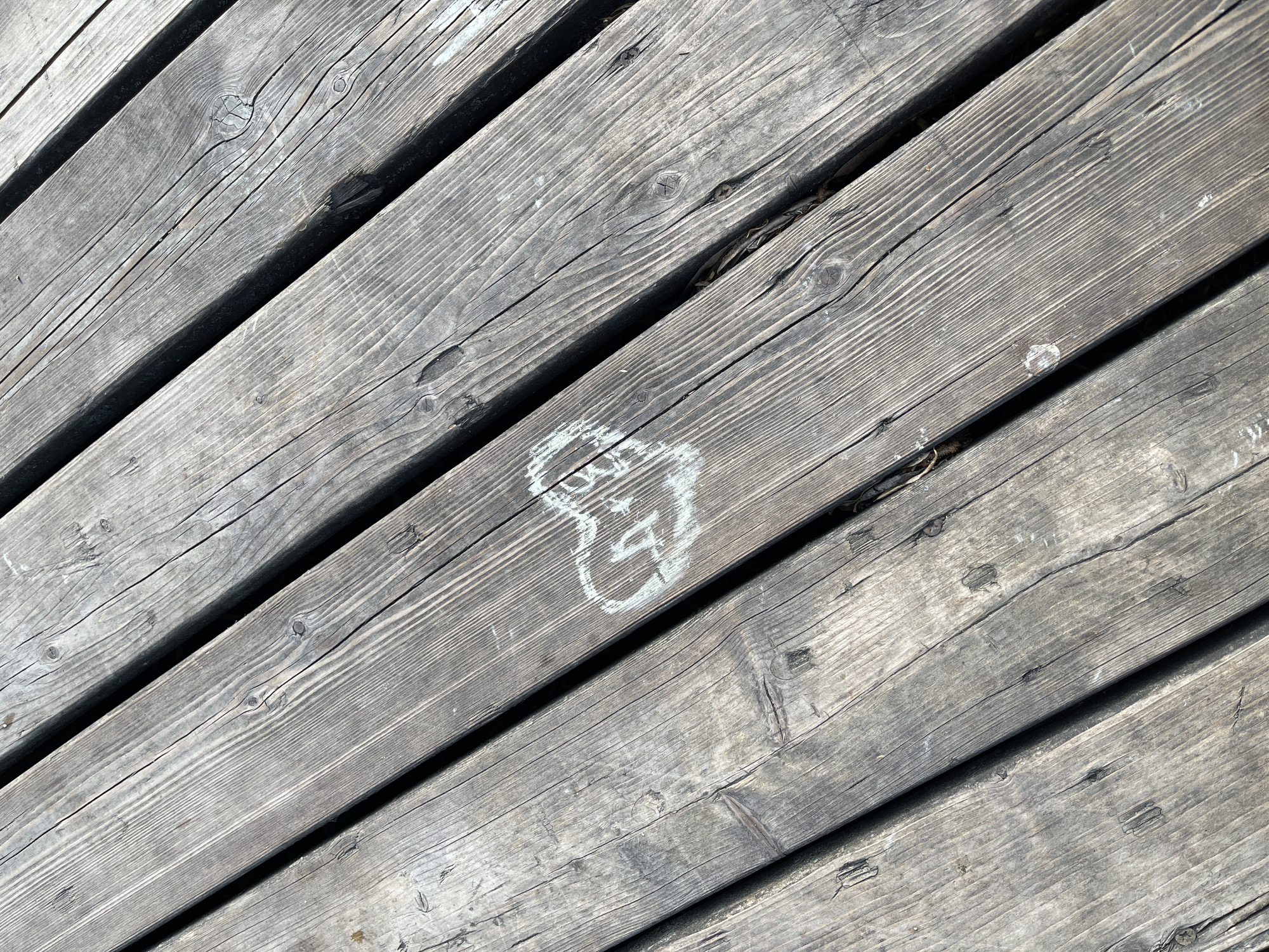 A close-up photograph depicts wooden decking planks arranged at a slight angle, revealing a weathered and aged surface. Visible wood grain and knots are present. A faint, roughly drawn "8" or infinity symbol is located towards the center of the image, appearing to be created with a light-colored substance, possibly chalk. The planks are separated by dark lines, indicating gaps between them. The overall tone is muted and gray. The image lacks any explicit indicators connecting it to Moscow, Russia. There is no indication of people, subjects, actions, or interactions.