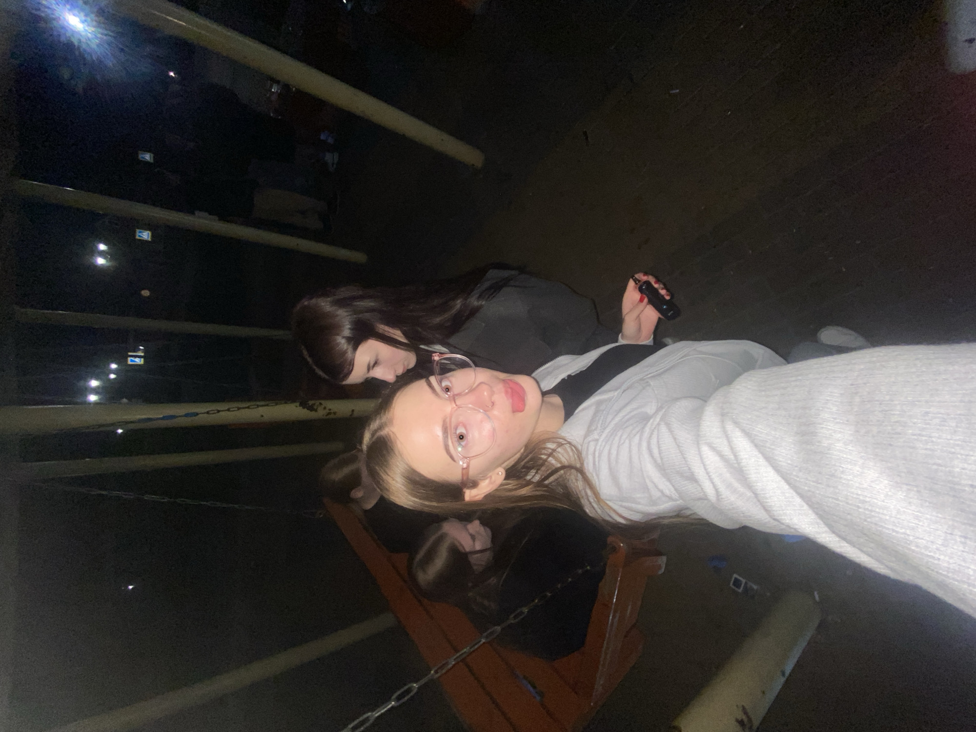 A low-light, grainy outdoor image depicts a group of young females at night in an unknown public area, likely a playground adjacent to a street in Unknown, Unknown. The scene is illuminated by distant streetlights and a direct flash source from the camera.
In the immediate foreground, a young female with light brown hair, wearing pink-framed eyeglasses and a light-colored ribbed top over a black shirt, is taking a selfie. She has a stud piercing on her left nostril and displays red nail polish on her right hand, which holds a small black electronic device resembling a vape pen. Her tongue is slightly extended, and her gaze is directed at the camera.
Behind and to her left, a wooden bench-style swing is visible, supported by four yellow-tan metal poles. Two females sit on the swing, both with dark hair and wearing dark clothing; one is mostly obscured, while the other faces away from the camera. A third female with long dark hair stands to the right of the swing, to the foreground subject's left, looking towards the left.
In the dimly lit background, multiple bright streetlights exhibit starburst patterns, and at least two blue and white road signs (possibly pedestrian crossing indicators) are visible. Several blurred figures are discernible further back. The ground is dark, appearing to be paved, with minor debris visible.