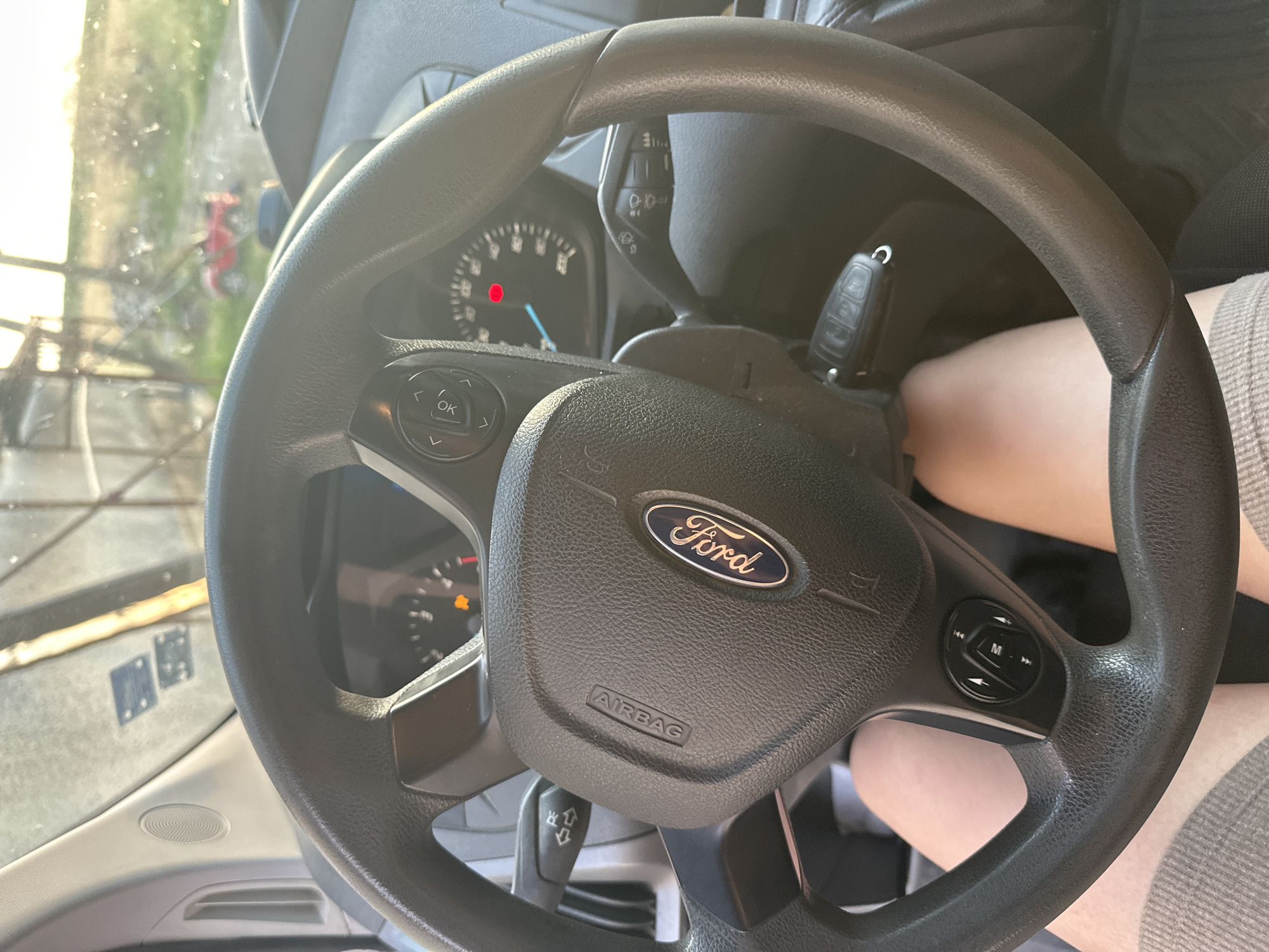 The media file is a close-up, interior shot of a Ford vehicle's steering wheel and dashboard. The steering wheel is black and features the Ford logo prominently in the center. Below the logo, the word "AIRBAG" is visible. On the left side of the steering wheel, there are buttons with directional arrows and an "OK" button. On the right side, there is a circular control with buttons labeled "M", and forward/backward arrows, likely for media controls. A car key is inserted into the ignition on the right side of the steering column.
The dashboard includes a speedometer with a red warning light illuminated and an orange indicator light. The windshield offers a blurred view of an outdoor scene, with greenery and a red vehicle visible in the distance. Sunlight streams in, suggesting it is daytime, and the weather appears clear. The overall scene depicts the interior of a vehicle, possibly a van or truck, ready for operation. No people are clearly visible, though a person's legs are partially visible beneath the steering wheel. The setting appears to be a parking area or roadside, given the view through the windshield. No specific text beyond the visible labels on the steering wheel and dashboard is discernible. The context provided indicates the location is Mlekarevo, Bulgaria.
