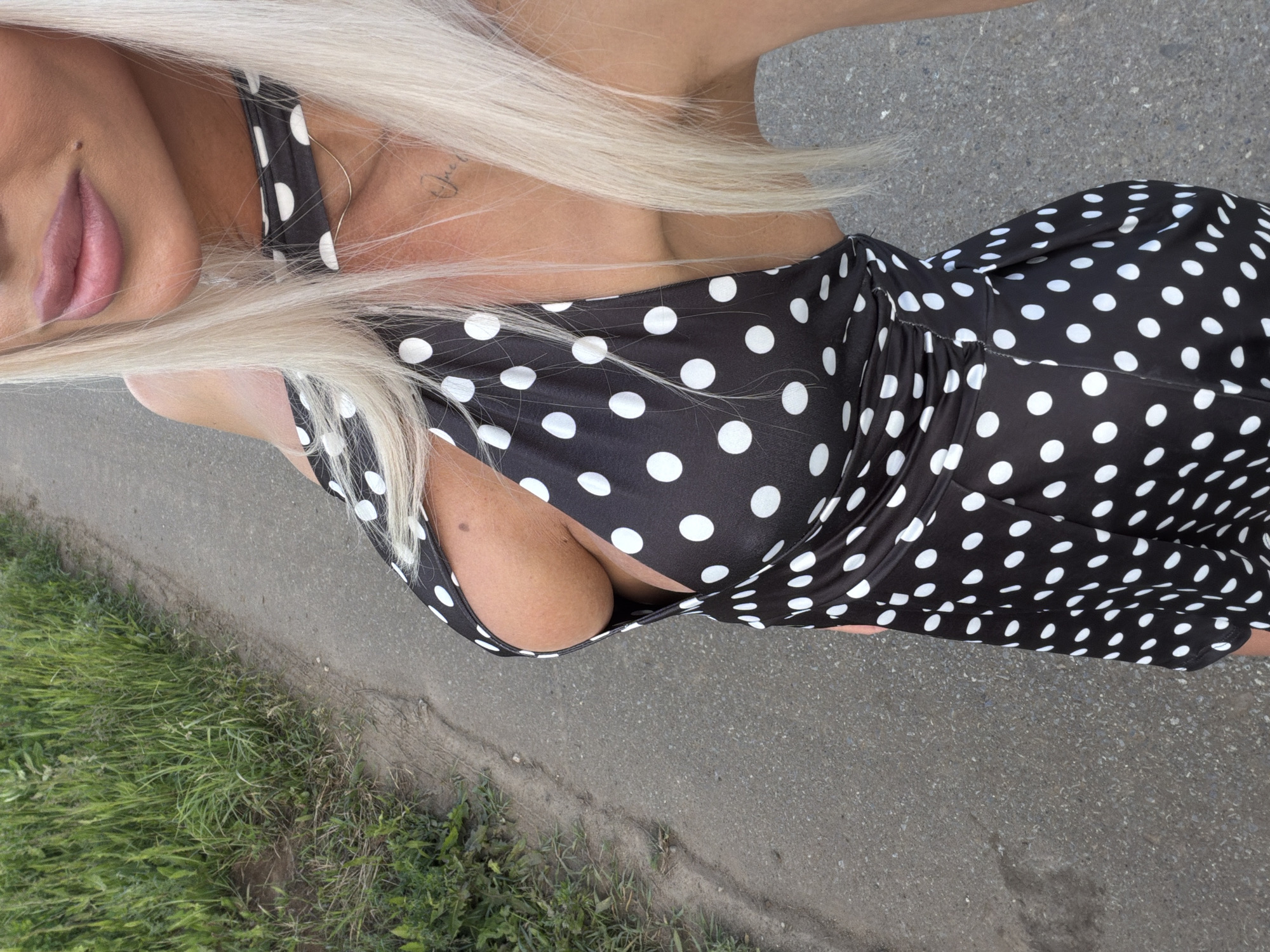 The image depicts a close-up, partial view of a person outdoors, rotated 90 degrees clockwise. The subject has long, platinum blonde hair and is wearing a black garment adorned with white polka dots, possibly a dress or a top with thin straps, which exposes cleavage. A golden hoop earring is visible on their right side, near the jawline. Their face is partially shown from the nose down, highlighting full, light pink lips. A small, cursive tattoo, which appears to read "Jack" or a similar name, is visible on their upper chest/collarbone area.
The setting is outdoors during daylight hours, characterized by clear ambient lighting. The person is positioned on a paved surface, likely asphalt or concrete, with visible aggregate. Alongside the paved area, a strip of green grass and various weeds grows, suggesting a roadside, pathway, or park environment. The photograph is taken from a slightly elevated perspective, looking down at the subject. The person appears to be casually posing for the camera. No other individuals or significant objects are present in the frame. The location context provided is Конь, Russia.