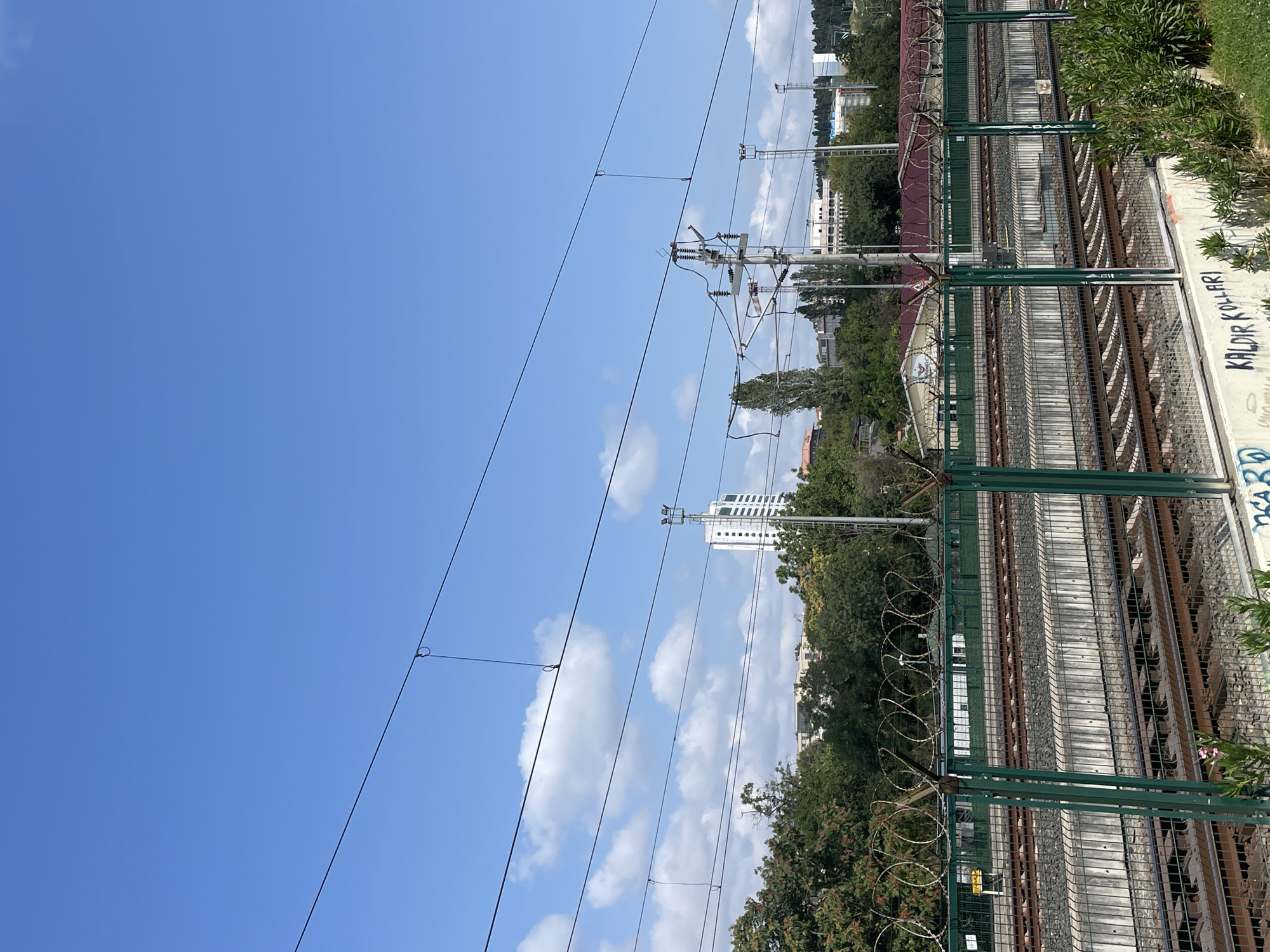 The image depicts a view of train tracks and overhead wires against a bright blue sky with scattered white clouds. A green metal fence with barbed wire runs across the foreground, separating the viewer from the train tracks. The tracks are laid out in parallel lines, with concrete barriers between them. Beyond the tracks, a line of trees and buildings is visible, including a tall white skyscraper and other structures. The scene appears to be in an urban or suburban area, likely in Kadıköy, Turkey, given the presence of train infrastructure and urban development. There are no people visible in the image. The overall impression is one of infrastructure and urban landscape under a clear sky.