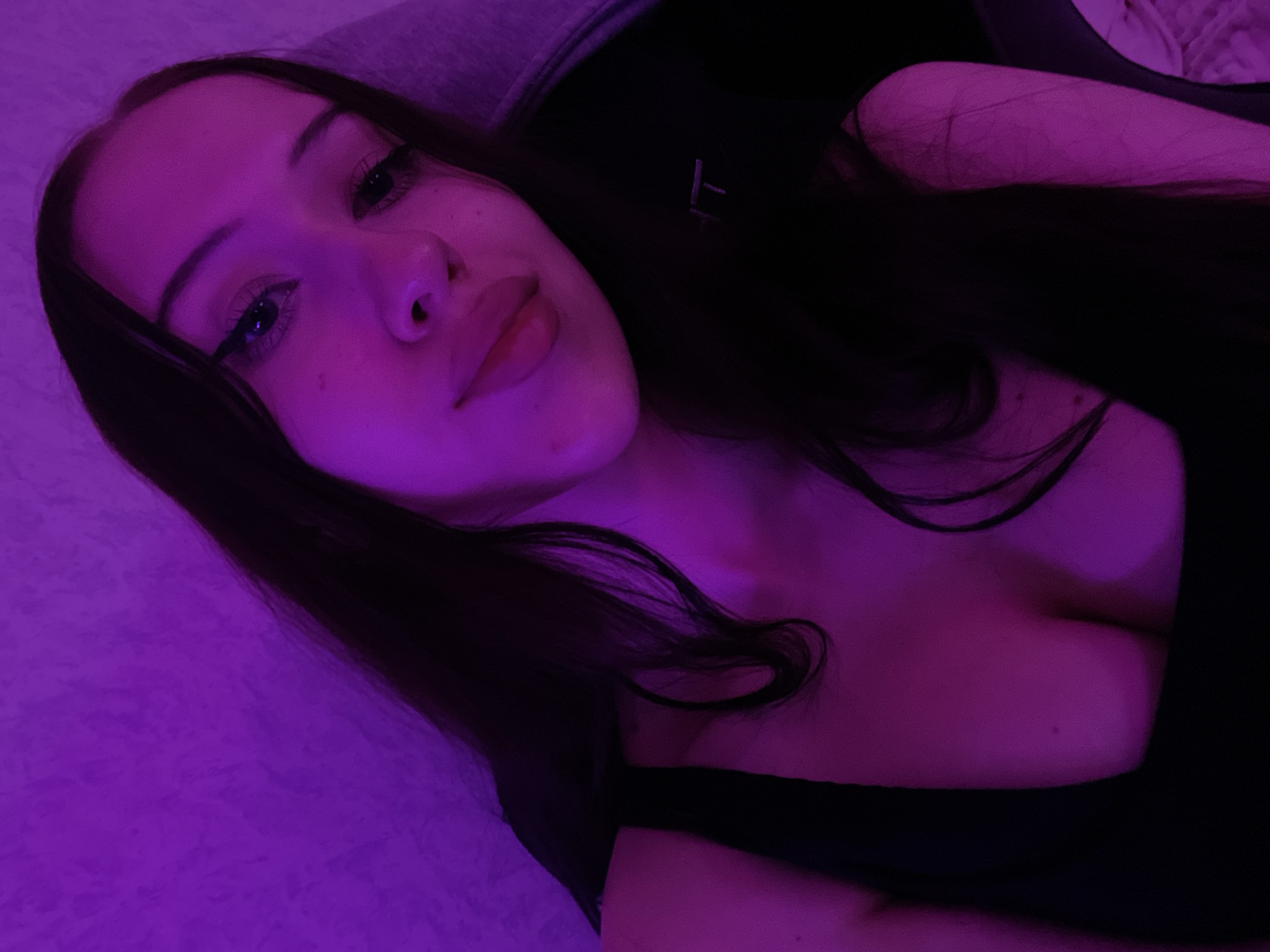 A young woman with long, dark hair and dark eyes, wearing a black top, poses for a selfie. The image is bathed in purple light, creating a moody atmosphere. The background appears to be a textured wall. The woman has dark eyeliner on her eyes and her lips are slightly parted. The scene is indoors, likely in a residential setting. There is no information in the image to suggest a location in Votkinsk, Russia.