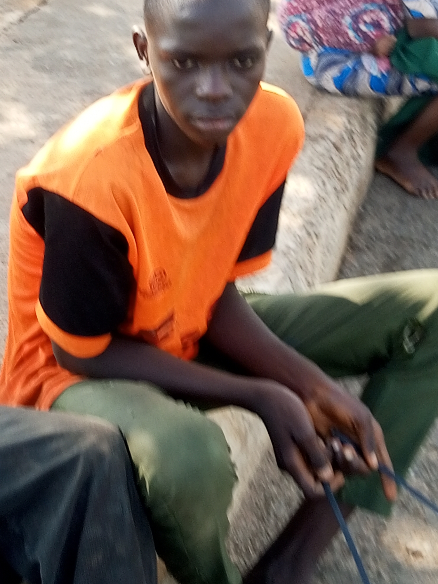 In Tempure, Nigeria, a young boy with dark skin and short hair is seated outdoors. He is wearing an orange t-shirt with black sleeves and green pants. His hands are clasped, manipulating a thin blue cord, possibly engaged in a task or game. The ground appears to be a mixture of sand and dirt. In the background, another person is partially visible, seated and wearing a patterned fabric. The lighting suggests it is daytime.