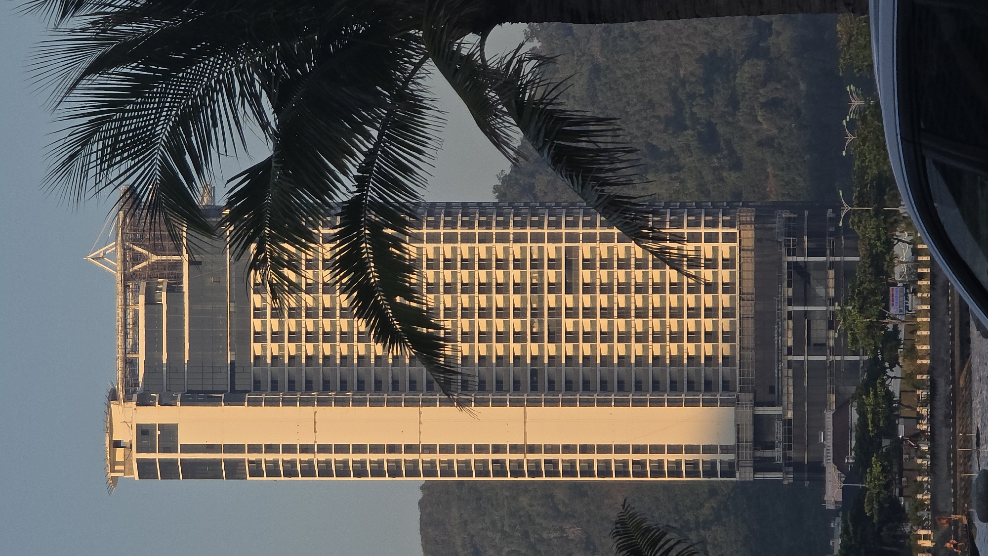 A tall, multi-story building under construction dominates the center of the image, illuminated by warm, golden light on its right facade, indicating either sunrise or sunset in Nha Trang, Vietnam. The structure exhibits numerous uniformly arranged windows or balcony openings across its main body. A prominent lighter-toned vertical section runs along its left side. The building's upper levels feature exposed structural elements and a distinctive triangular framework on the rooftop, signifying ongoing construction. Dark construction netting is visible on some of the lower floors. A large, dark green palm frond occupies the right foreground, partially obscuring the building. The roofline and side window of a dark-colored vehicle are visible at the bottom right. In the midground, across a body of water, a coastal road with streetlights and sparse foliage is discernible, with indistinct figures near the waterline on the far left. Hazy, tree-covered mountains rise in the background behind the building, under a clear, light blue sky.