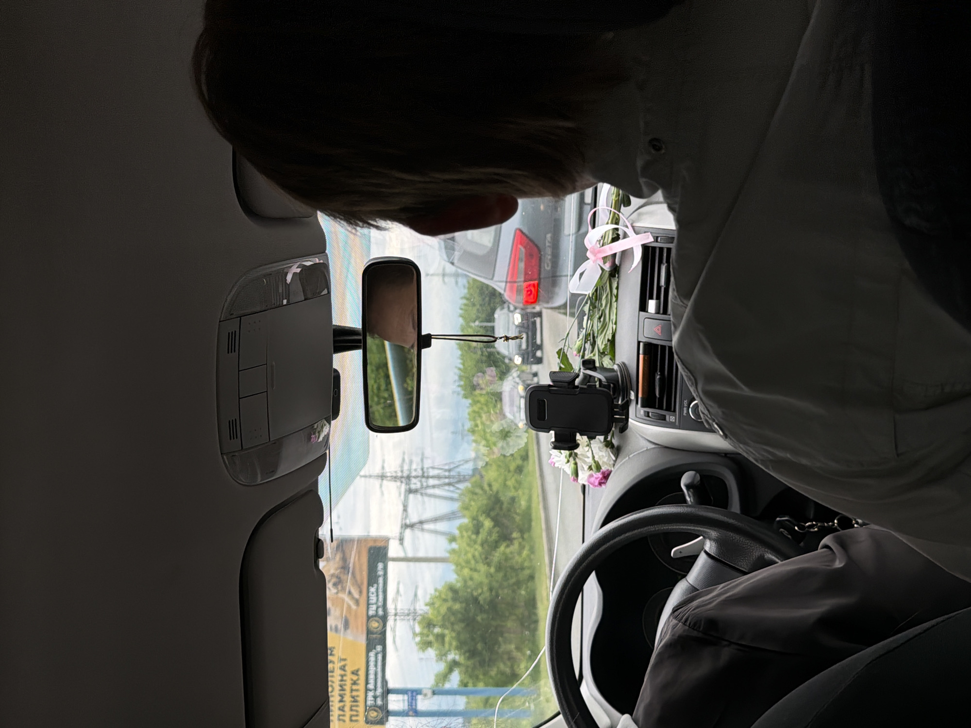 The image shows the interior of a car, seemingly taken from the passenger seat. The scene is oriented at an angle.
Inside the car: A person with short, light-colored hair leans towards the center console. The car has a rear-view mirror, a phone mount, and a bouquet of flowers adorned with a pink ribbon on the dashboard. A steering wheel is visible on the right side.
Outside the car (as seen through the windshield): Several cars are visible on the road ahead, including a gray Hyundai Creta. High-voltage power lines are seen against a cloudy sky. Roadside advertising signs with text in Russian and images of construction materials are also visible.
Location: Based on the Cyrillic text on the advertising signs, the image is likely taken in or near the city of Chaikovsky, Russia. Specifically, the billboard shown has text that says "ЛАМИНАТ ПЛИТКА", the Russian words for laminate and tile. One billboard has the address for TPK Akvarel', which is located in Chaikovsky, Russia.