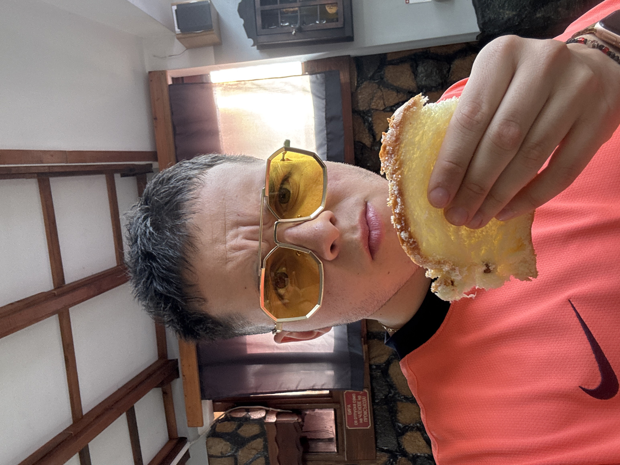 A sideways shot depicts a man wearing yellow-tinted glasses and a salmon-colored Nike shirt, holding a piece of cake. The setting appears to be inside a building with exposed wooden beams and a stone wall. A sign in Cyrillic script is partially visible, suggesting the location is in a Bulgarian-speaking area, possibly near Kostinbrod, Bulgaria. A television is also visible.