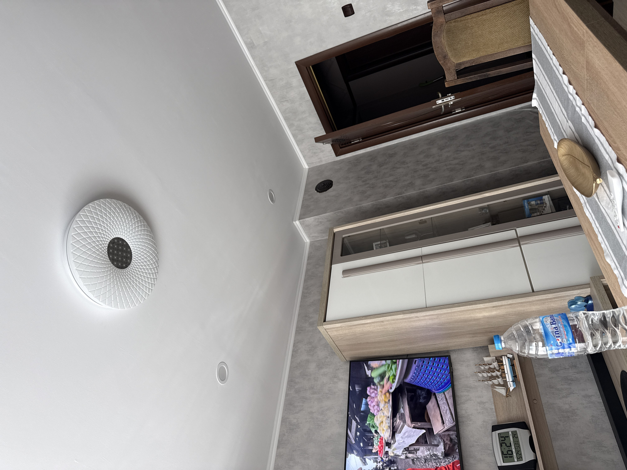 The image depicts an interior scene in Lovech, Bulgaria. It shows a white wall with a decorative circular ceiling light fixture, a recessed light, and a dark opening in the ceiling, possibly for ventilation or maintenance. Below, a wooden and white storage unit is partially visible, holding books and other items. A television displaying a marketplace scene is seen beneath the storage unit, next to a small shelf with a water bottle, a model sailboat, and a digital clock displaying "16:24." A wooden table with a striped runner and a golden decorative bowl sits to the right. The overall impression is of a modern, tidy living space.