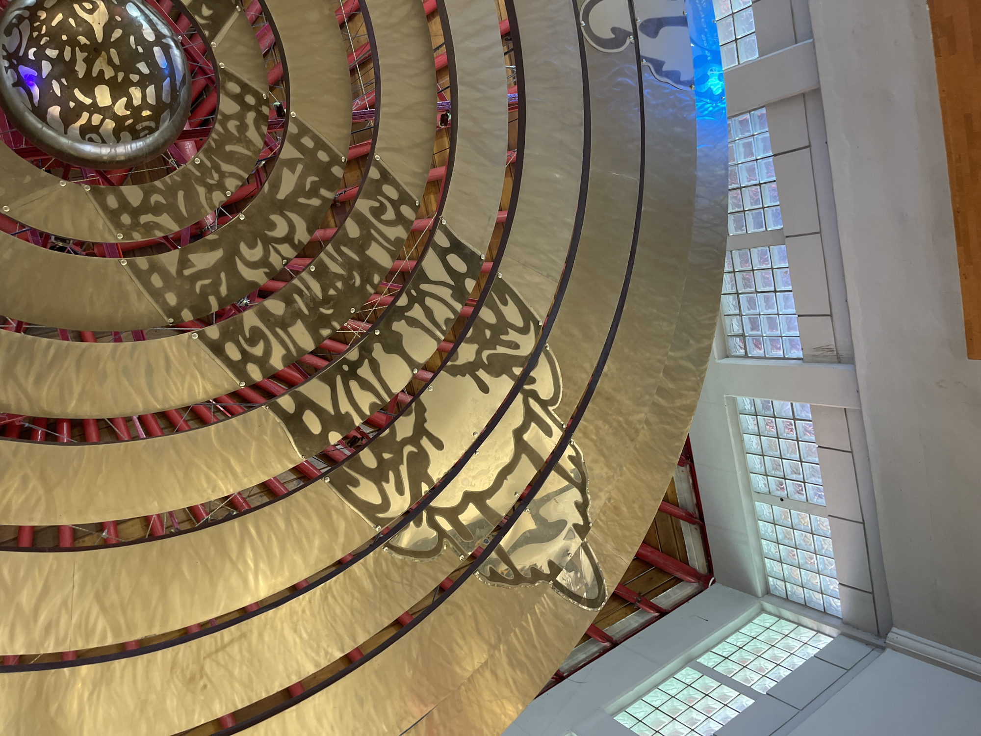 This image depicts the interior of a building, likely a religious or cultural space, in Kuta, Indonesia. The most prominent feature is a large, tiered ceiling made of reflective golden material arranged in concentric circles. These circular tiers are supported by red beams and are adorned with intricate, dark patterned cutouts on some sections, resembling organic or abstract designs. The center of the ceiling has a circular element with colored lights, reflecting onto the metallic surfaces.
Below the ceiling, on the left side of the image, are several windows constructed with frosted glass blocks arranged in a grid pattern. These windows allow diffused natural light to enter the space. The walls appear to be painted white. The architectural style suggests a blend of modern and potentially traditional influences, given the decorative ceiling and the use of glass block windows. There are no people visible in the image. The overall impression is one of a well-lit, possibly serene interior space with a striking, artful ceiling.
