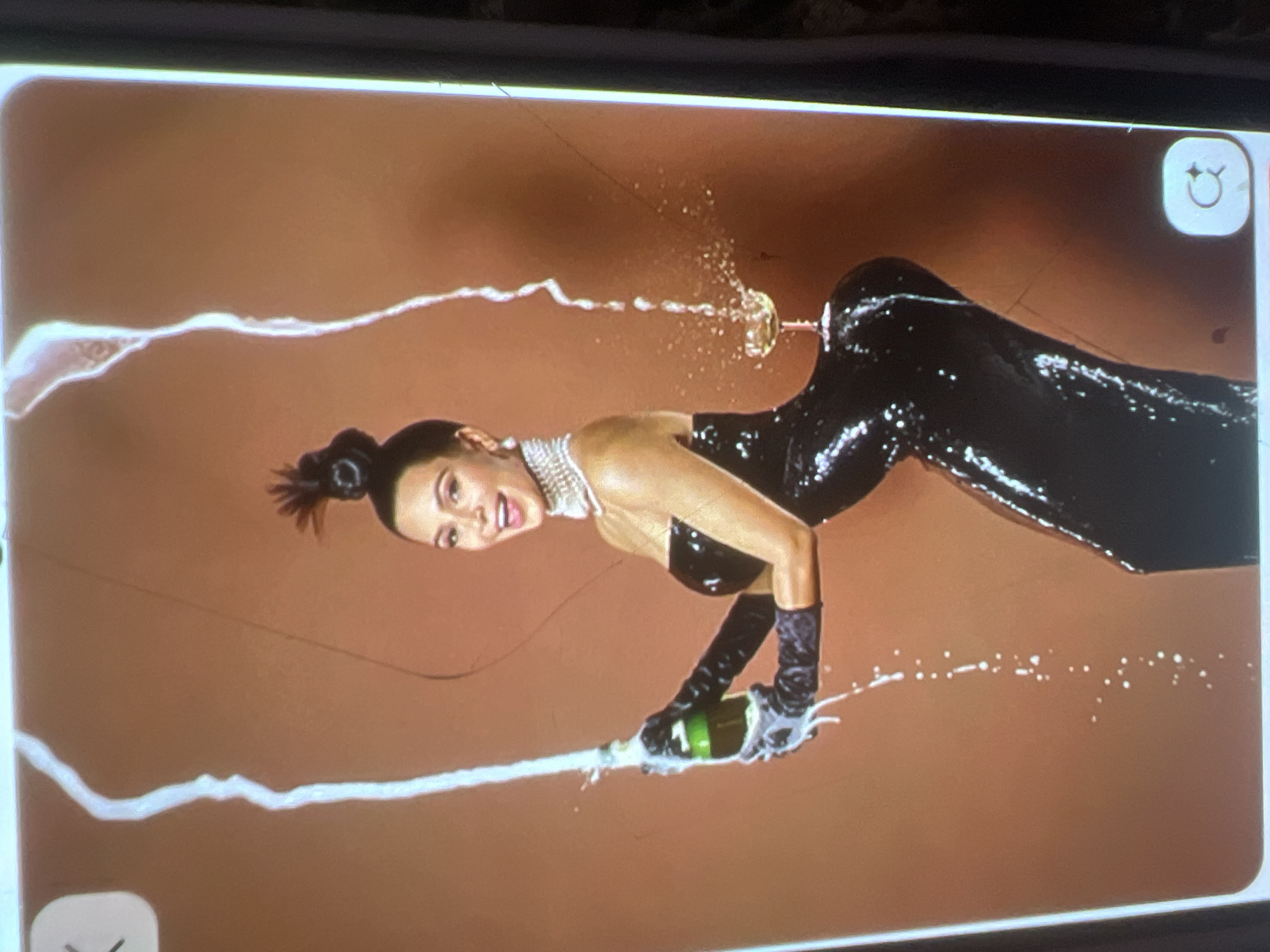 The image shows a woman in a black, form-fitting, sequined dress and long black gloves. She is holding a green bottle of champagne with a green label and the cork has been removed. Champagne is being sprayed from the bottle in a large arc. She is also wearing a pearl necklace and pearl earrings. Her hair is pulled up into a high bun with a few strands falling around her face. She is smiling and looking towards the viewer. To her right, a champagne coupe glass is precariously balanced on her hip. The background is a solid, reddish-brown color. There are also some white streaks that appear to be part of the champagne spray or a digital effect. The image is a vertical photograph, likely taken indoors given the controlled lighting and background. There is no discernible connection to Kara-Dıykan, Kyrgyzstan, or any specific urban environment; the setting is abstract.