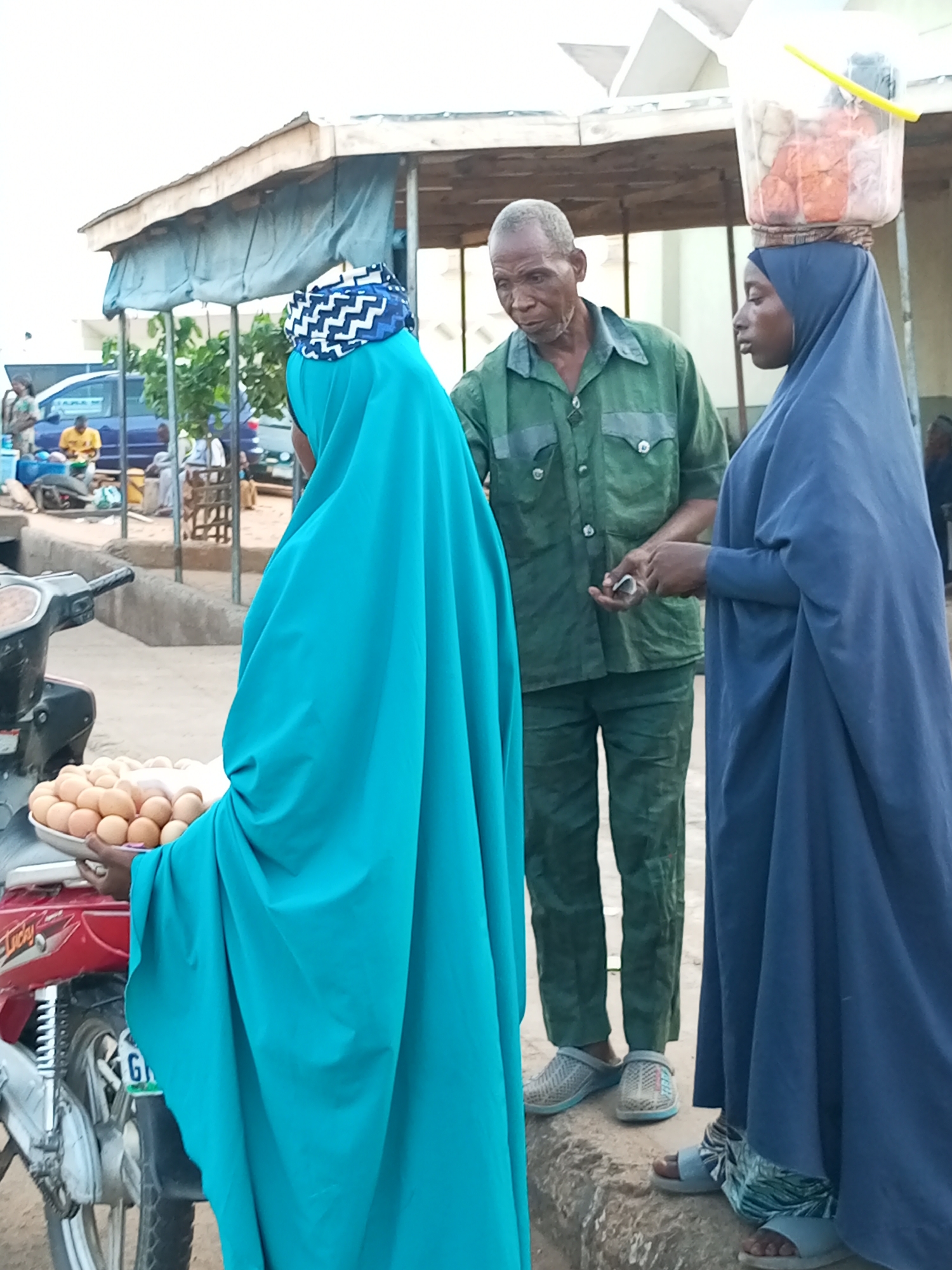 In Gombe, Nigeria, an outdoor market scene unfolds. Three individuals are visible, with two women and one man in the foreground. A woman on the left, wearing a bright teal hijab and matching abaya, holds a tray of eggs. To her right, a man in a green shirt and trousers appears to be making a transaction with another woman. This second woman, on the far right, is dressed in a dark blue hijab and abaya, and has a bucket of items balanced on her head. Behind them, a red motorcycle with a license plate is parked. The environment suggests a bustling street or market with a stall-like structure providing shade in the background.