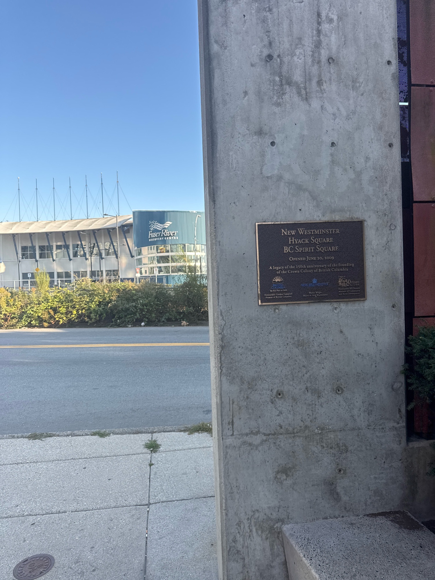 The image shows a bronze plaque affixed to a concrete pillar. The plaque reads: "NEW WESTMINSTER HYACK SQUARE BC SPIRIT SQUARE OPENED JUNE 20, 2009 A legacy of the 150th anniversary of the founding of the Crown Colony of British Columbia." Below this text are logos for the province of British Columbia, the City of New Westminster, and a "BC 50 YEARS" logo. Also on the plaque are the names of officials, including "Honourable Gordon Campbell Premier of British Columbia" and "Wayne Wright Mayor of New Westminster."
In the background, a modern building with a curved, light-colored facade and large windows is visible. A blue sign on this building reads "FRASER RIVER DISCOVERY CENTRE." The building is surrounded by lush green bushes and trees. A paved road with double yellow lines runs in front of the bushes, and a concrete sidewalk is visible in the foreground. The sky is clear and blue. The overall scene depicts an outdoor public space in New Westminster, Canada.
