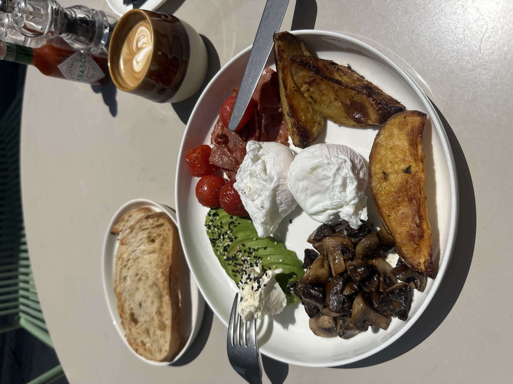 The image shows a table setting in a restaurant, presumably located in Abu Dhabi, United Arab Emirates. A plate with a breakfast meal takes center stage, containing fried potatoes, bacon, cherry tomatoes, two poached eggs, avocado slices sprinkled with sesame seeds, cream cheese, and sautéed mushrooms. A knife rests on the plate, and a fork is positioned nearby. A separate small oval dish holds two slices of bread. A coffee cup with latte art, a Tabasco sauce bottle, and a salt or pepper grinder are also visible on the table. The table's surface appears to be a light grey color. Part of a green chair is visible in the lower-left corner of the frame.