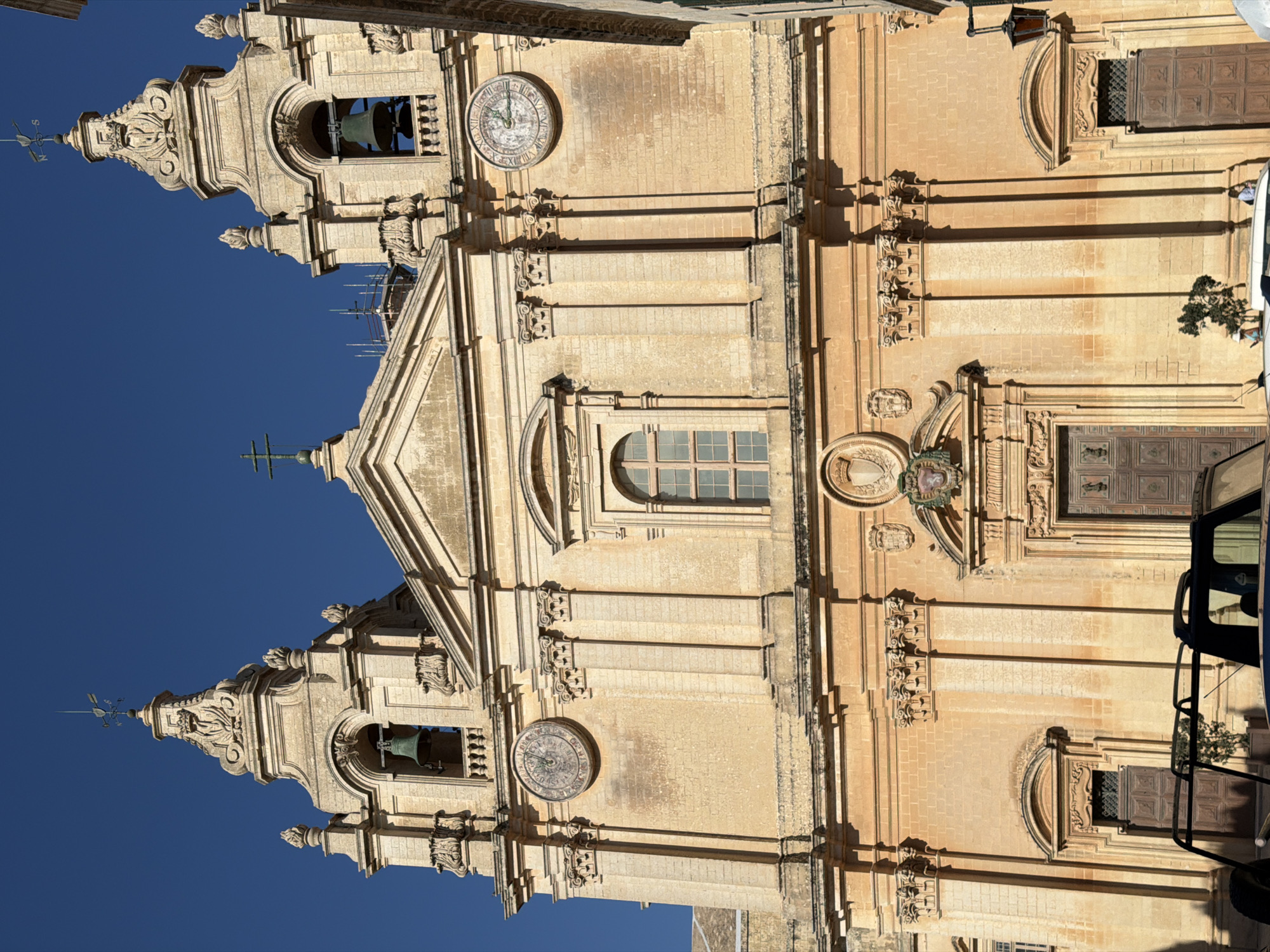 The image shows a close-up view of the facade of a historic stone building, likely a church, with elaborate architectural details. The building is constructed from light-colored stone and features a symmetrical design with two bell towers, one on each side of the central structure. Each tower has an arched opening containing a large bell and is topped with ornate decorative elements, including sculptures and a weather vane.
In the center of the facade, above a triangular pediment, is a large circular clock with Roman numerals. Further down, there are several rectangular windows with grid patterns and arched doorways. The building's facade is richly decorated with pilasters, cornices, and decorative carvings. Shadows cast by the sun indicate it is daytime, likely mid-morning or early afternoon, with clear blue skies.
The image is oriented vertically, with the building's facade filling most of the frame. The lower right corner of the image shows the side of a dark-colored vehicle, possibly a van or SUV, with a black metal cage or rack attached to it. A small green tree is visible behind the vehicle, and a few people are seen near the building and the vehicle. The overall impression is one of a grand, ancient structure bathed in sunlight.
The location context provided is Mdina, Malta, which aligns with the architectural style visible in the image. There is no discernible text visible in the image.
