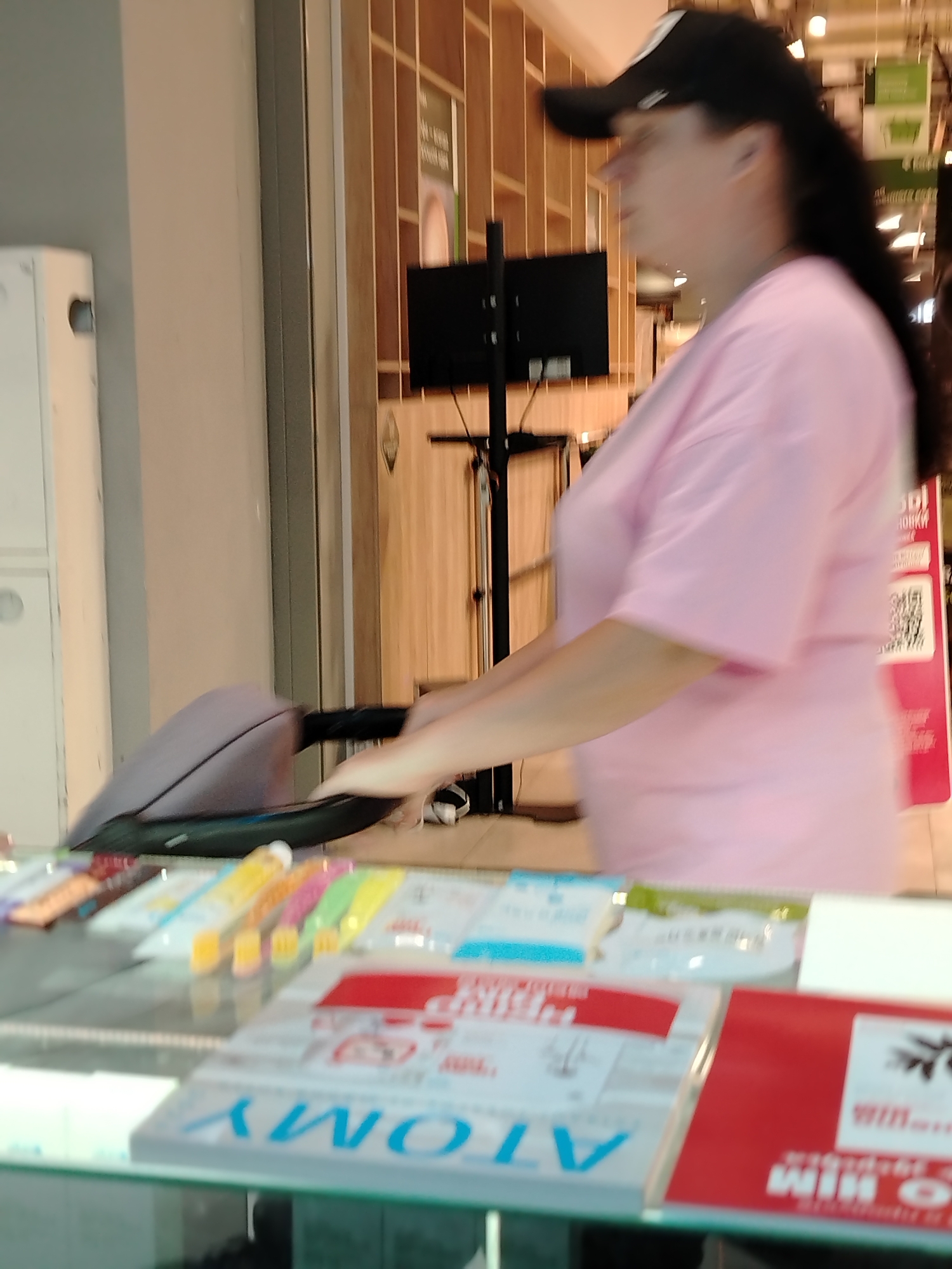 The image shows a woman with long black hair and a black baseball cap, wearing a pink t-shirt, standing next to a display of goods. The display features a stack of "ATOMY" branded products, along with other items that appear to be packaged goods. In the background, there is a display screen on a stand, and a wall with wooden paneling. There is likely an air conditioning unit or other type of mechanical unit in the left background as well. The location appears to be a commercial space, possibly in Izhevsk, Russia, given the brand name appearing on some items. The woman is looking to the right, and her hand seems to be near the handle of a stroller.