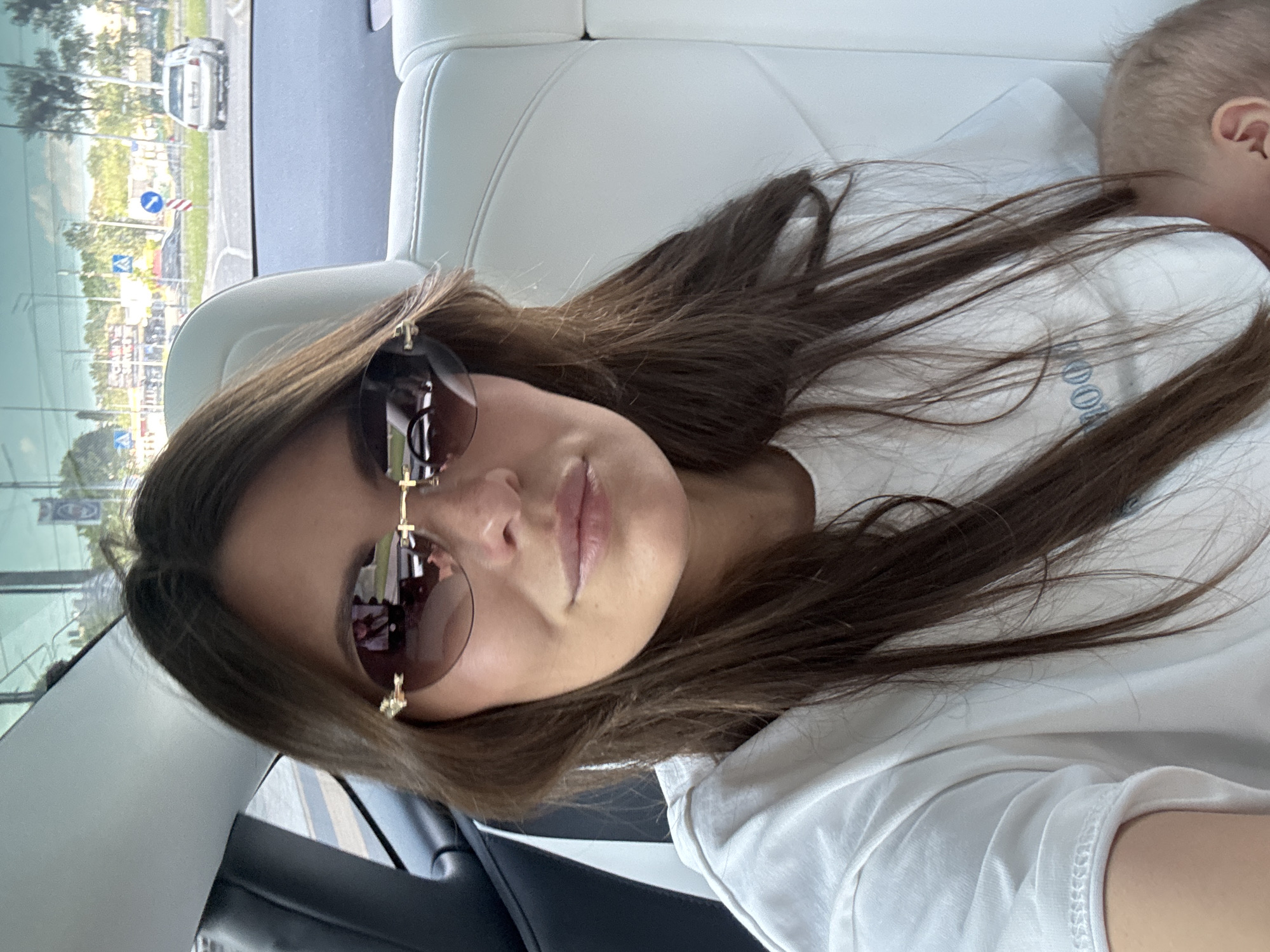 The image captures a woman and a baby inside a vehicle during daytime. The woman, positioned in the foreground, has long, dark brown hair and is wearing a white t-shirt and sunglasses with distinctive gold cross-shaped embellishments on the frames. Her expression appears relaxed, with lips slightly parted. On her white shirt, faint blue text, possibly spelling "MOON," is partially visible.
To the woman's right, the back of a baby's or toddler's head with light-colored hair and an ear is partially visible, suggesting the child is resting against the vehicle's white seat or headrest. The interior of the vehicle features white, possibly leather or faux leather, seating.
Through the window in the background, a bright outdoor scene is visible, indicative of daylight hours. It shows a road with a white car in the distance, green trees, and some structures. A blue circular traffic sign is also discernible, indicating an urban or suburban setting consistent with Sofiivska Borshchahivka, Ukraine. The overall activity depicted is a casual moment, possibly a selfie, during travel.