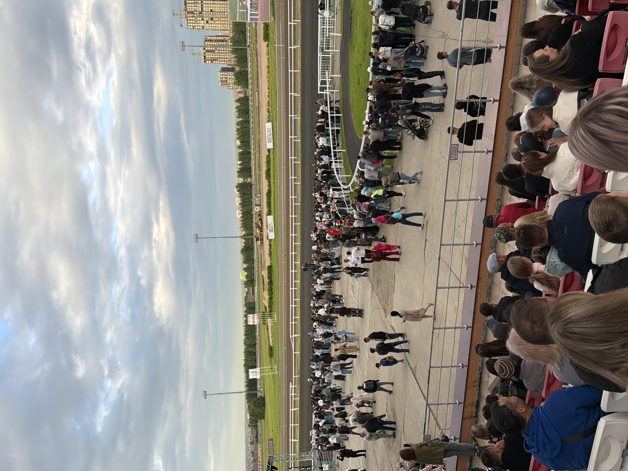 The image depicts an outdoor event at a racetrack or stadium, set against a cloudy sky. In the foreground, spectators are seated in rows of red and white stadium seats, facing away from the camera. Beyond the seating area, a large paved area is filled with a crowd of people milling about. This area is adjacent to a dirt racetrack bordered by white fencing and green grass. In the background, a line of trees separates the track from a cityscape featuring several tall, modern buildings under construction, indicated by visible cranes. The overall scene suggests a public gathering or sporting event.
