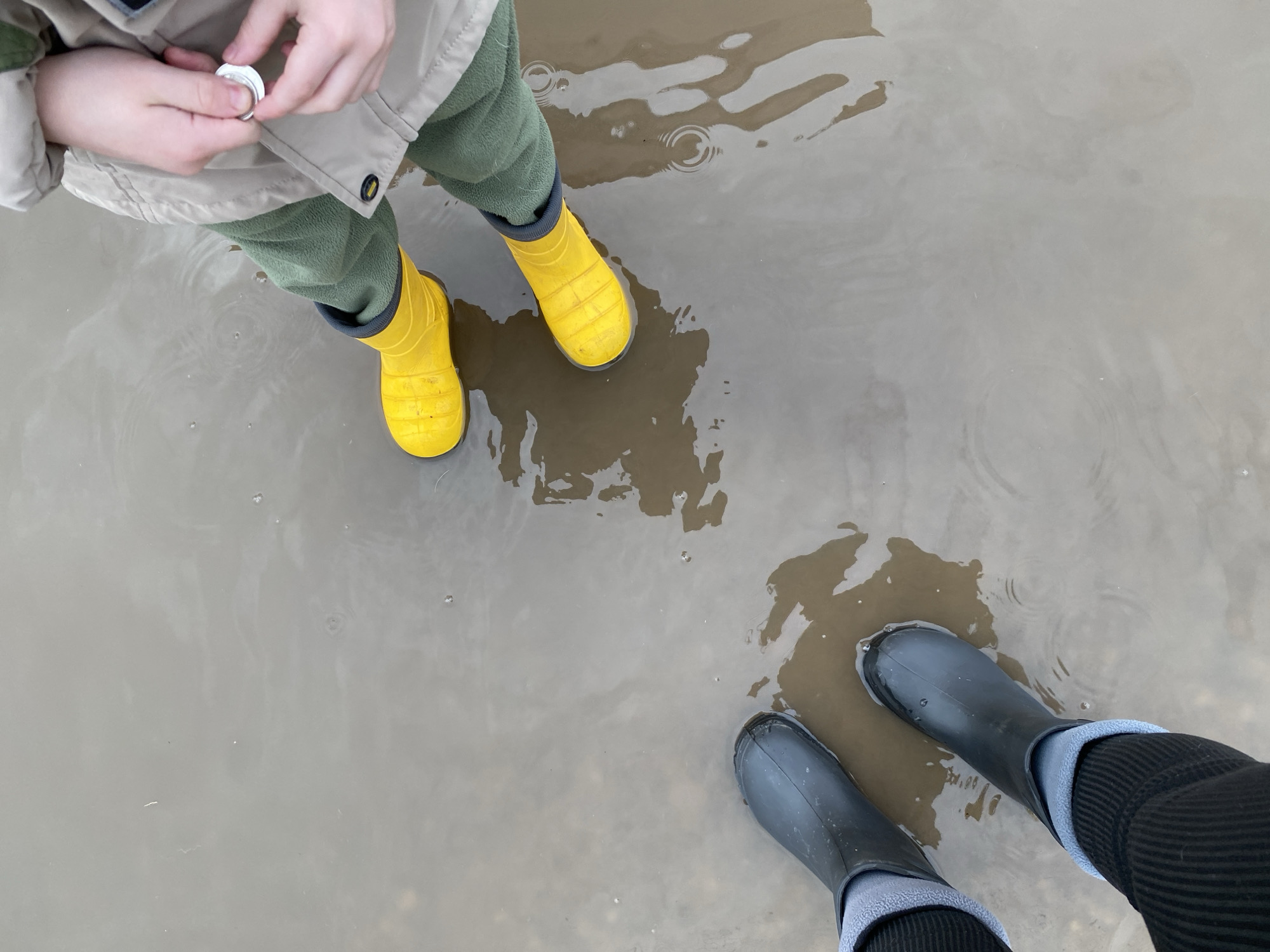 The image shows two people standing in a shallow pool of muddy water. One person is wearing yellow rubber boots, green pants, and a beige jacket, holding a small white object in their hands. The other person is wearing dark grey rubber boots, black pants, and grey socks. The location is Noviy, Russia. The setting is outdoors, and the ground is wet.