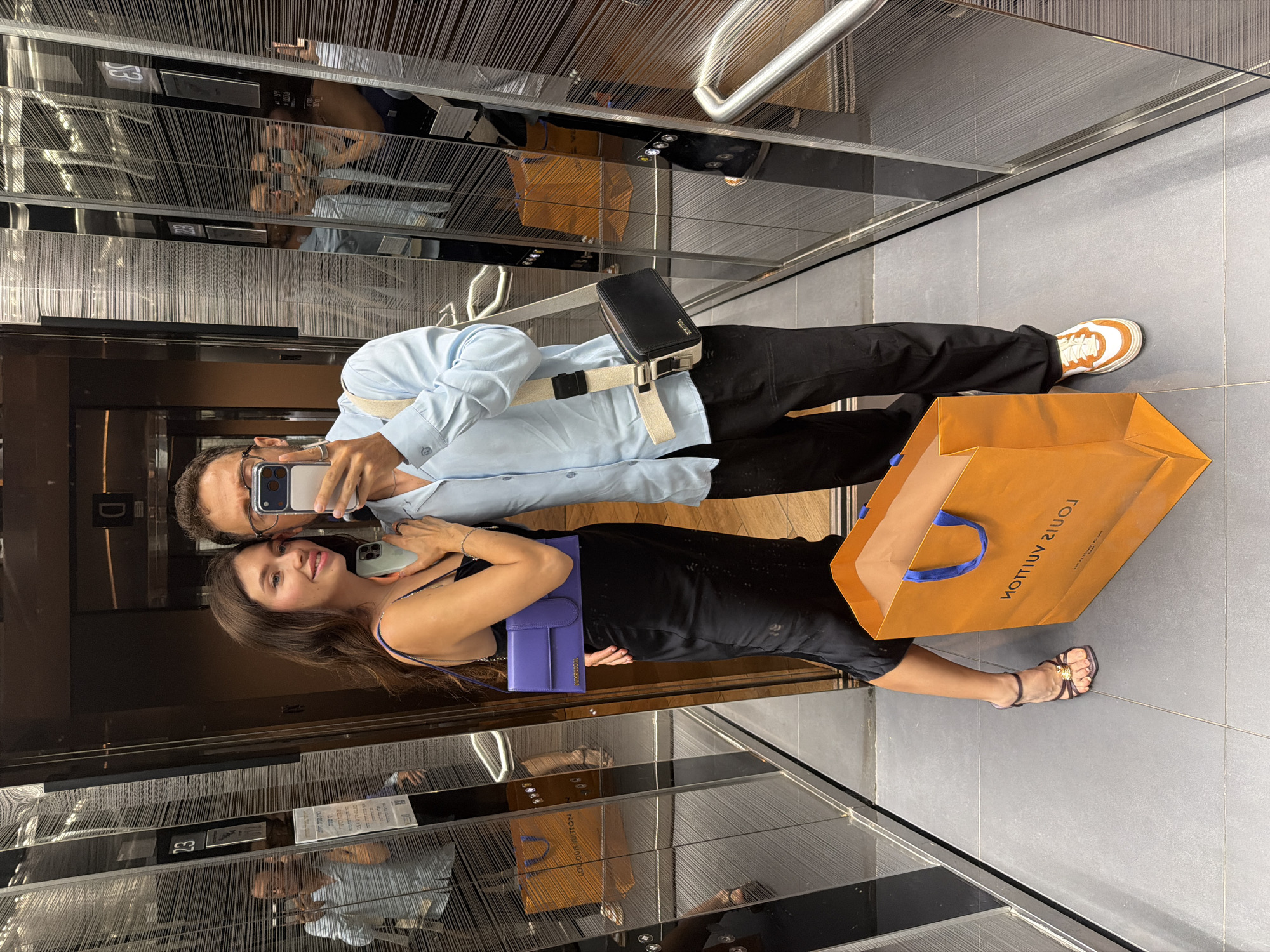 A young couple, a man and a woman, are taking a selfie in a modern elevator with mirrored walls. The woman, on the left, wears a black slip dress and a purple shoulder bag. The man, on the right, is dressed in a light blue collared shirt and black trousers, and carries a black crossbody bag. Both are wearing glasses. In front of them, on the floor, are two shopping bags, one orange and one brown. The elevator's interior features vertical striped paneling on the walls. The scene is set within a building in Tel-Aviv, Israel.