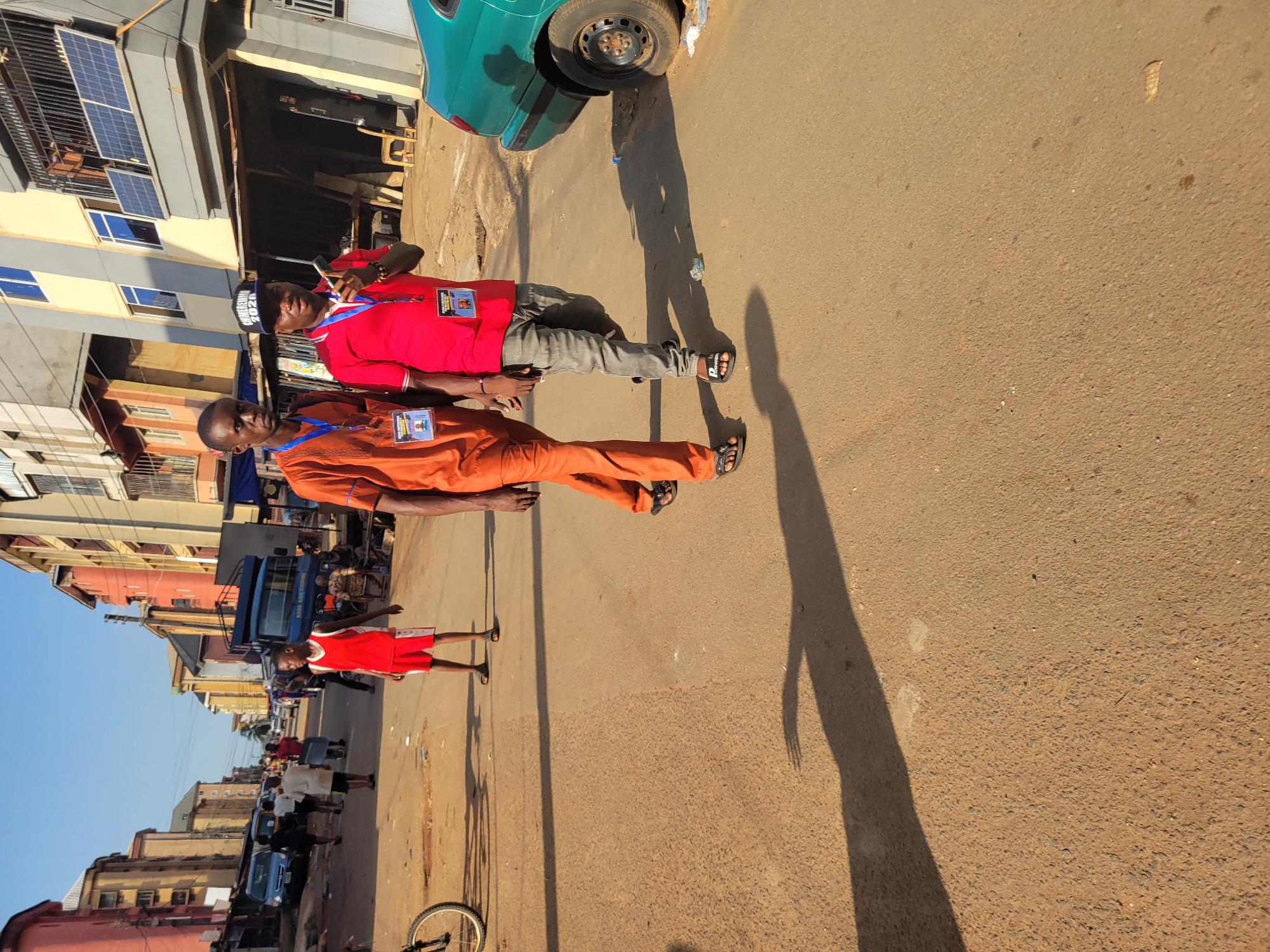 Two men are walking down a dirt road in Onitsha, Nigeria. The man on the left is wearing an orange robe and a lanyard with a badge. The man on the right is wearing a red shirt, gray pants, and a black baseball cap with "UNIMERERENA 2020" printed on it. He is holding a walkie-talkie to his ear. The road is lined with buildings and a blue truck. A green car is parked on the right side of the road. The sun is casting long shadows on the road.