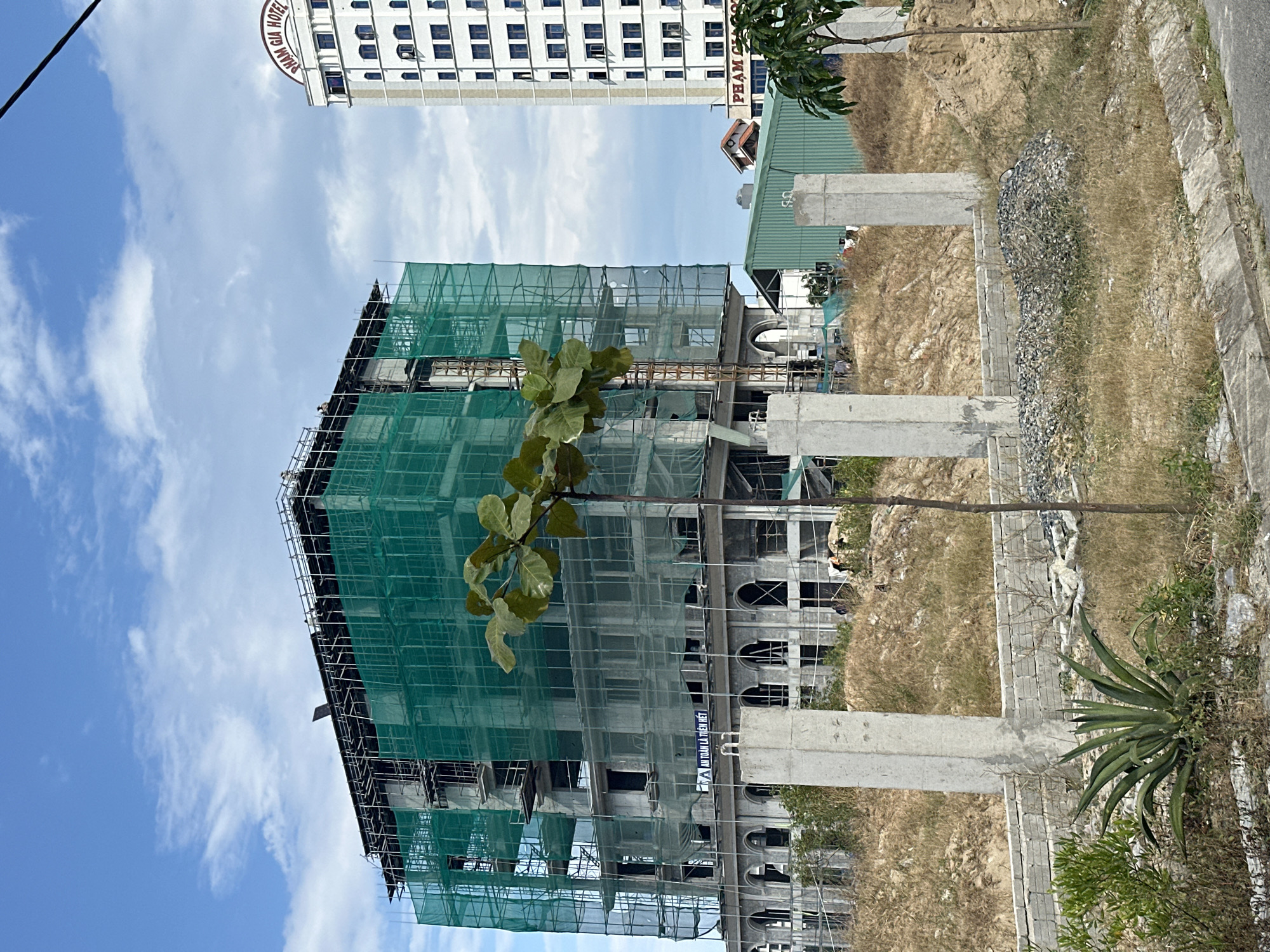The image shows an unfinished building under construction in Đà Nẵng, Vietnam. The building is covered in green netting and scaffolding. A second building, the Pham Gia Hotel, can be seen in the background. In the foreground, there are concrete pillars and dry vegetation. The sky is partly cloudy.