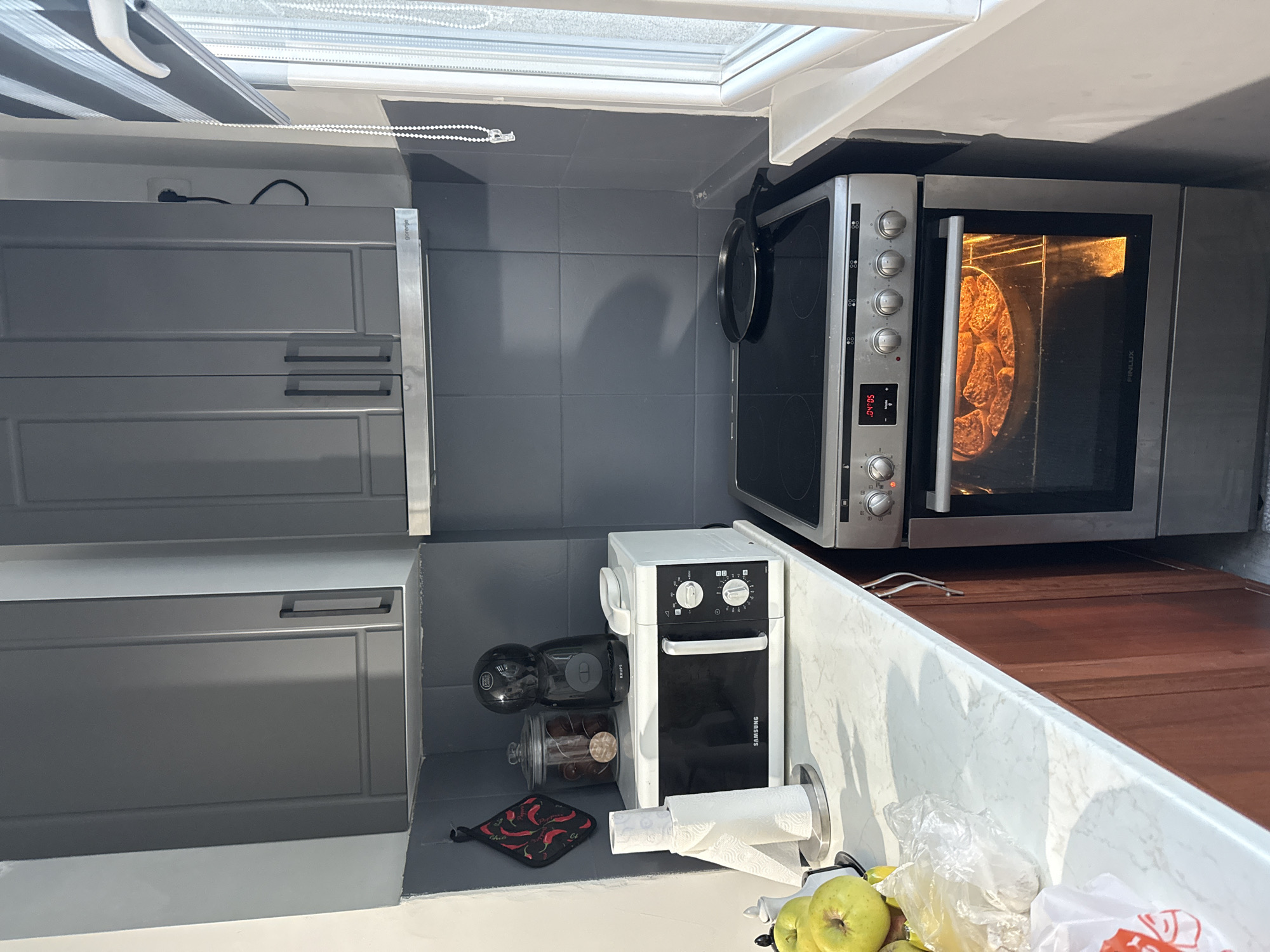 The image shows a kitchen interior, likely in Stara Zagora, Bulgaria, with gray cabinets on the left and an oven on the right. Inside the oven, a pizza is being baked. A small Samsung oven is situated under the larger oven next to a coffee maker. A roll of paper towels is also visible near the small oven, along with green apples and a bag on the counter. A chili patterned potholder sits nearby.